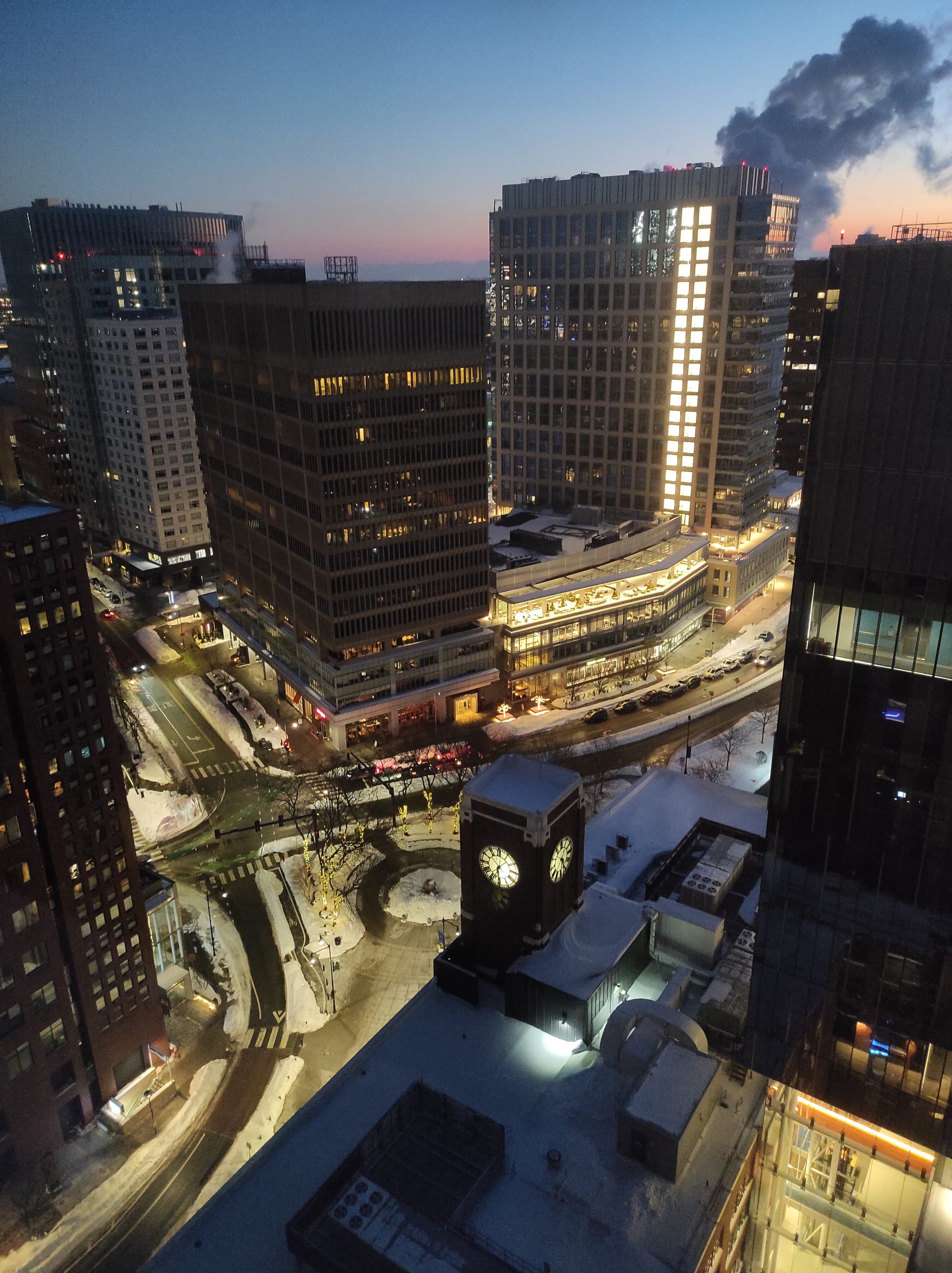 Ein Stadtbild in der Abenddämmerung mit schneebedeckten Straßen, hohen modernen Gebäuden und einem Uhrturm im Vordergrund. Einige Fenster sind hell erleuchtet, und Straßenlaternen beleuchten die verschneiten Straßen darunter. Von einem entfernten Dach steigt Dampf auf.