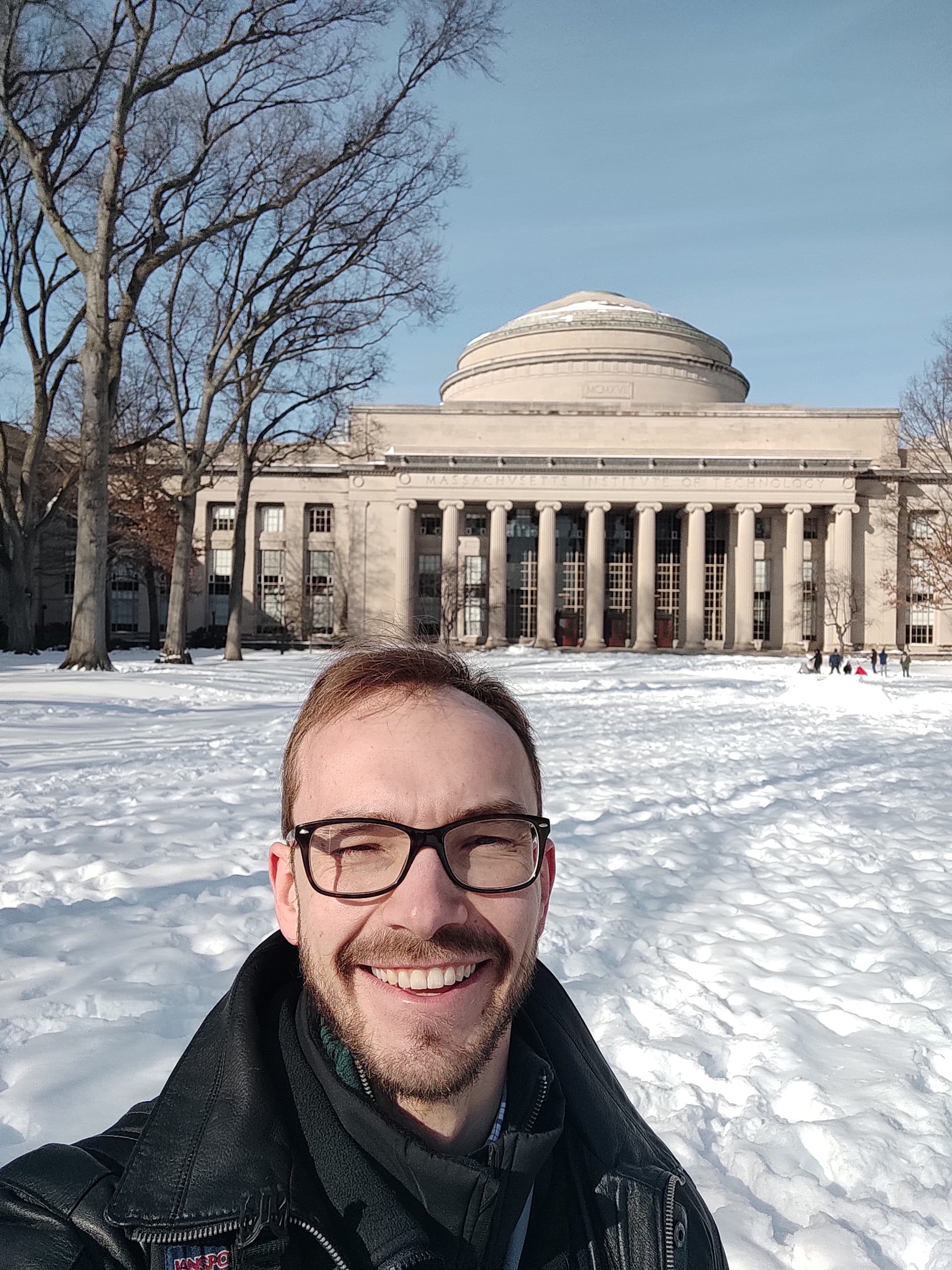 Ein Mann mit Brille und Bart lächelt für ein Selfie vor einem großen, kuppelförmigen Gebäude mit Säulen, umgeben von schneebedecktem Boden und kahlen Bäumen an einem sonnigen Tag.