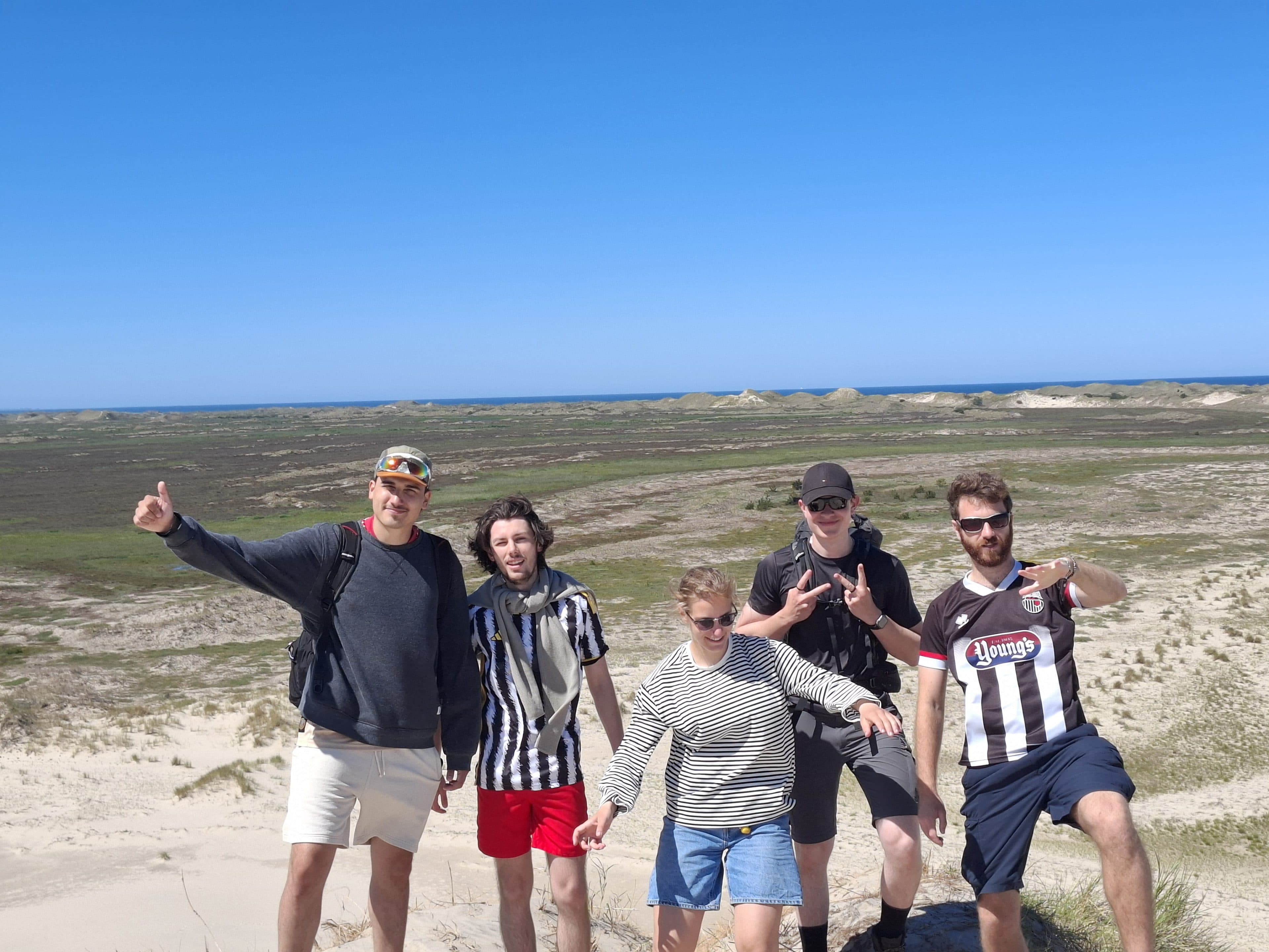 Fünf Männer stehen auf sandigen Dünen, im Hintergrund sind grünes Gras und ein blauer Himmel zu sehen. Sie posieren begeistert, einige zeigen den Daumen nach oben oder machen das Peace-Zeichen; sie tragen legere Sommerkleidung.