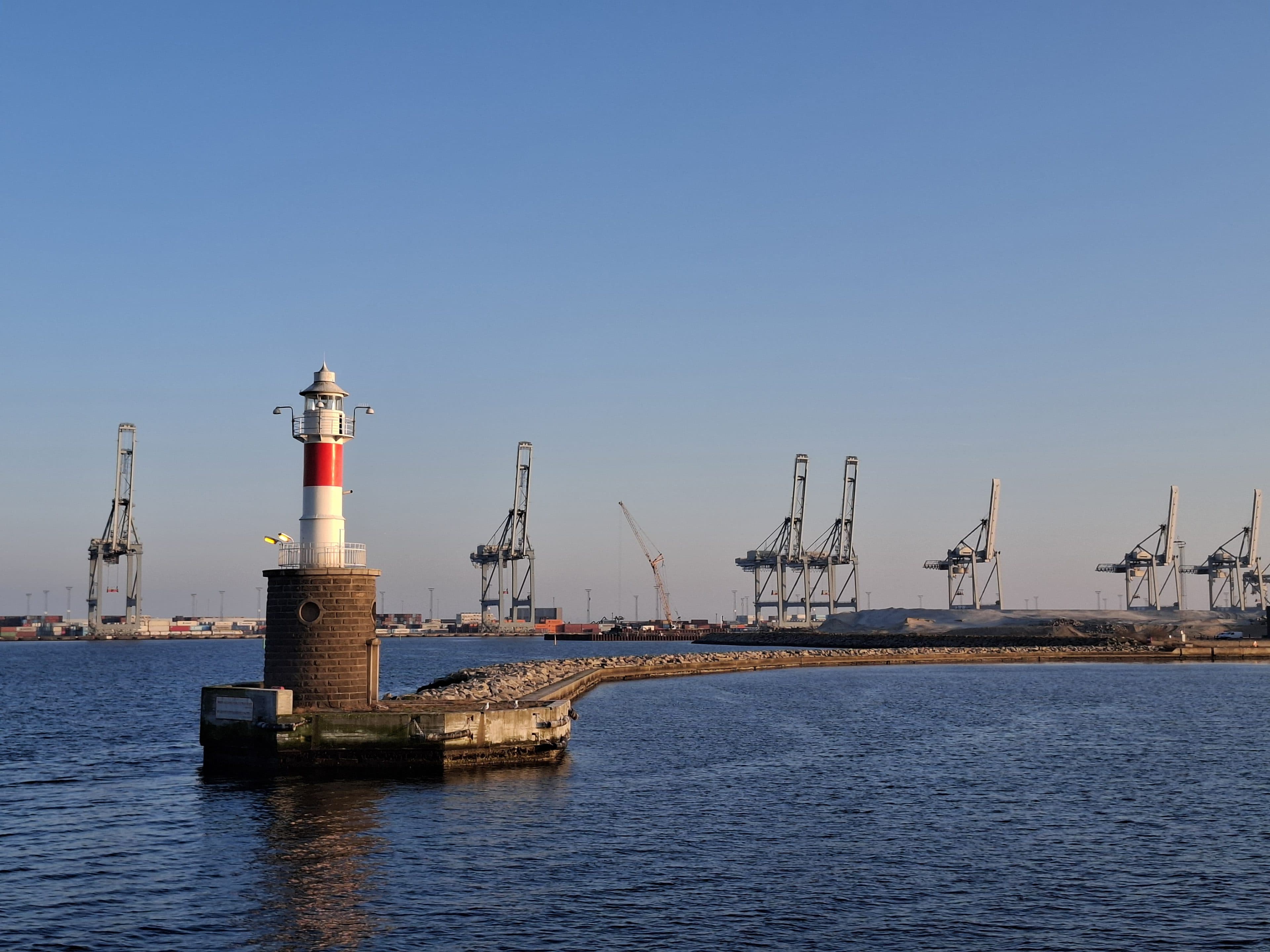 Am Ende eines geschwungenen Steinpiers steht ein kleiner, rot-weiß gestreifter Leuchtturm, im Vordergrund das ruhige blaue Wasser und am fernen Ufer unter einem klaren Himmel große Industriekräne.