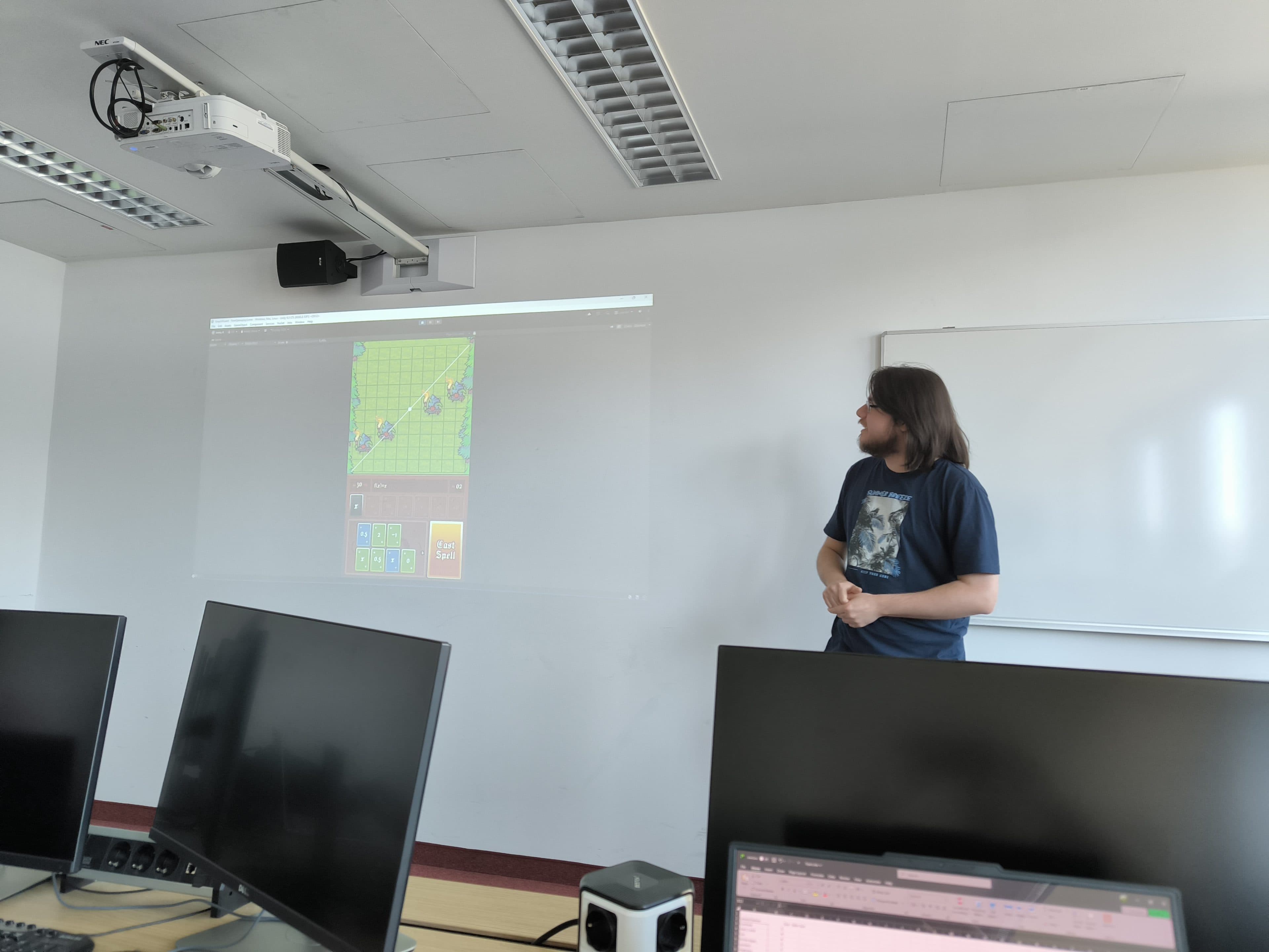 Eine Person steht vor einer Projektionsfläche, auf der ein buntes Gitter-Spiel zu sehen ist, in einem Klassenzimmer mit mehreren Computermonitoren und einer Whiteboard-Tafel.