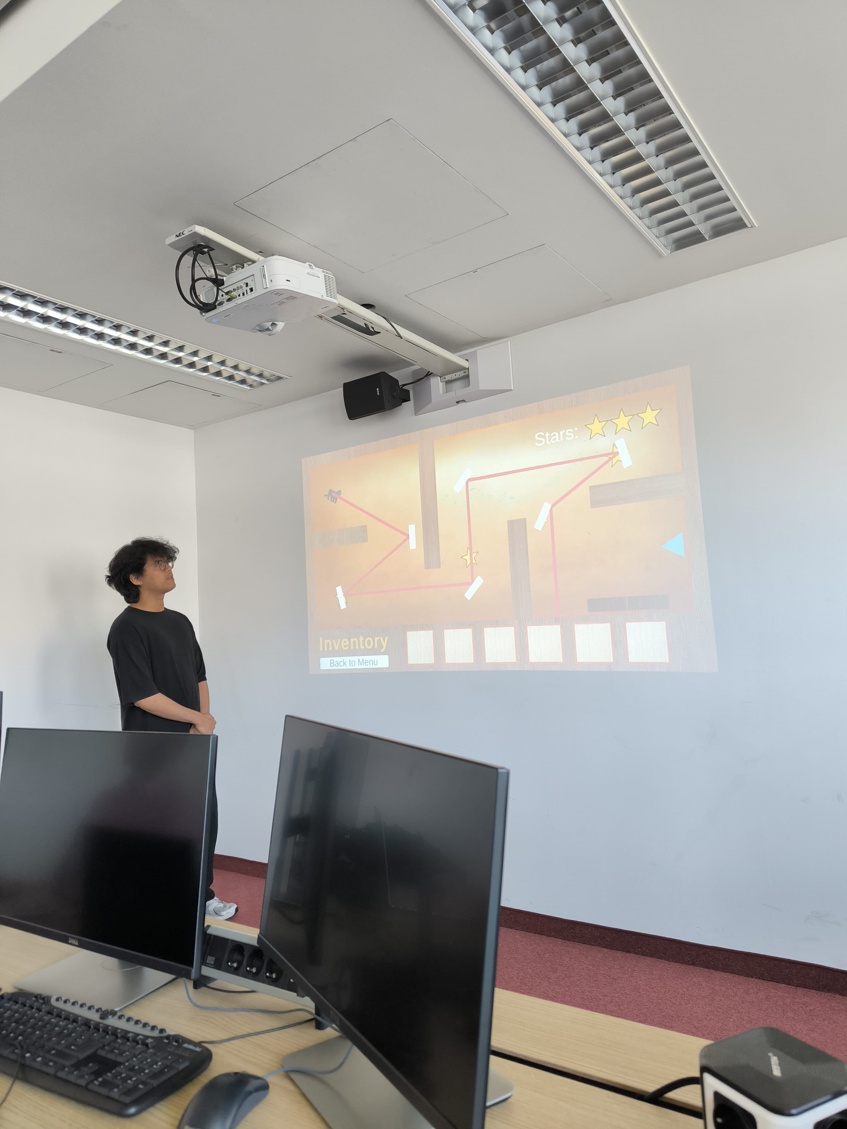 Eine Person steht in einem Klassenzimmer neben einer Leinwand, auf der ein Videospiel mit drei Sternen gezeigt wird. Im Vordergrund sind Computermonitore und Schreibtische zu sehen.