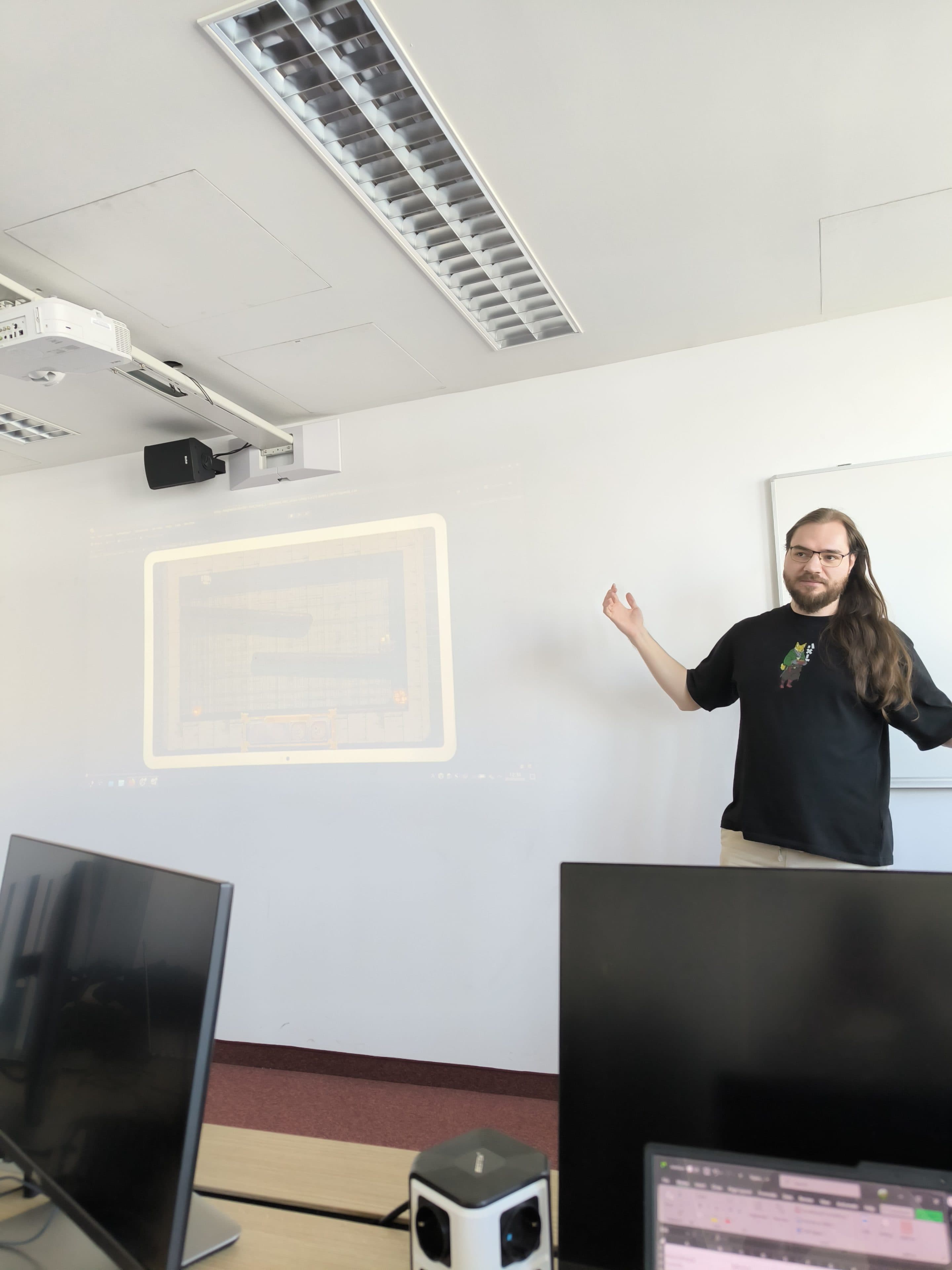 Eine Person mit langen Haaren steht vor einer Weißwandtafel und präsentiert ein projiziertes Bild eines Rasters oder eines Spiels auf einer Leinwand in einem Klassenzimmer, in dessen Vordergrund sich Computer auf den Tischen befinden.