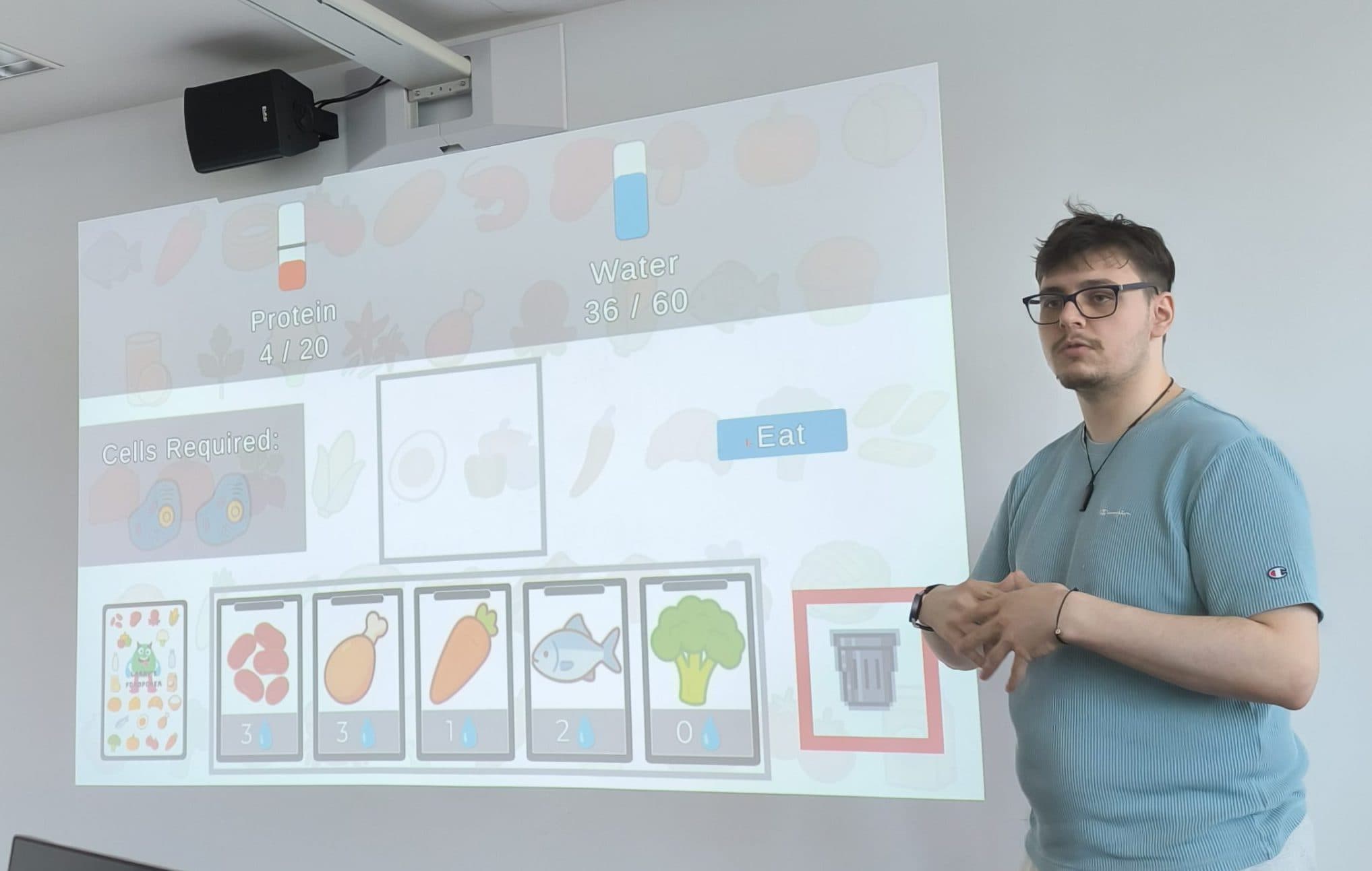 Eine Person in einem blauen Hemd steht in einem Klassenzimmer neben einer Projektionsfläche, auf der verschiedene Symbole und das Wort „Eat“ zu sehen sind. Im Vordergrund sind mehrere Computermonitore zu sehen.