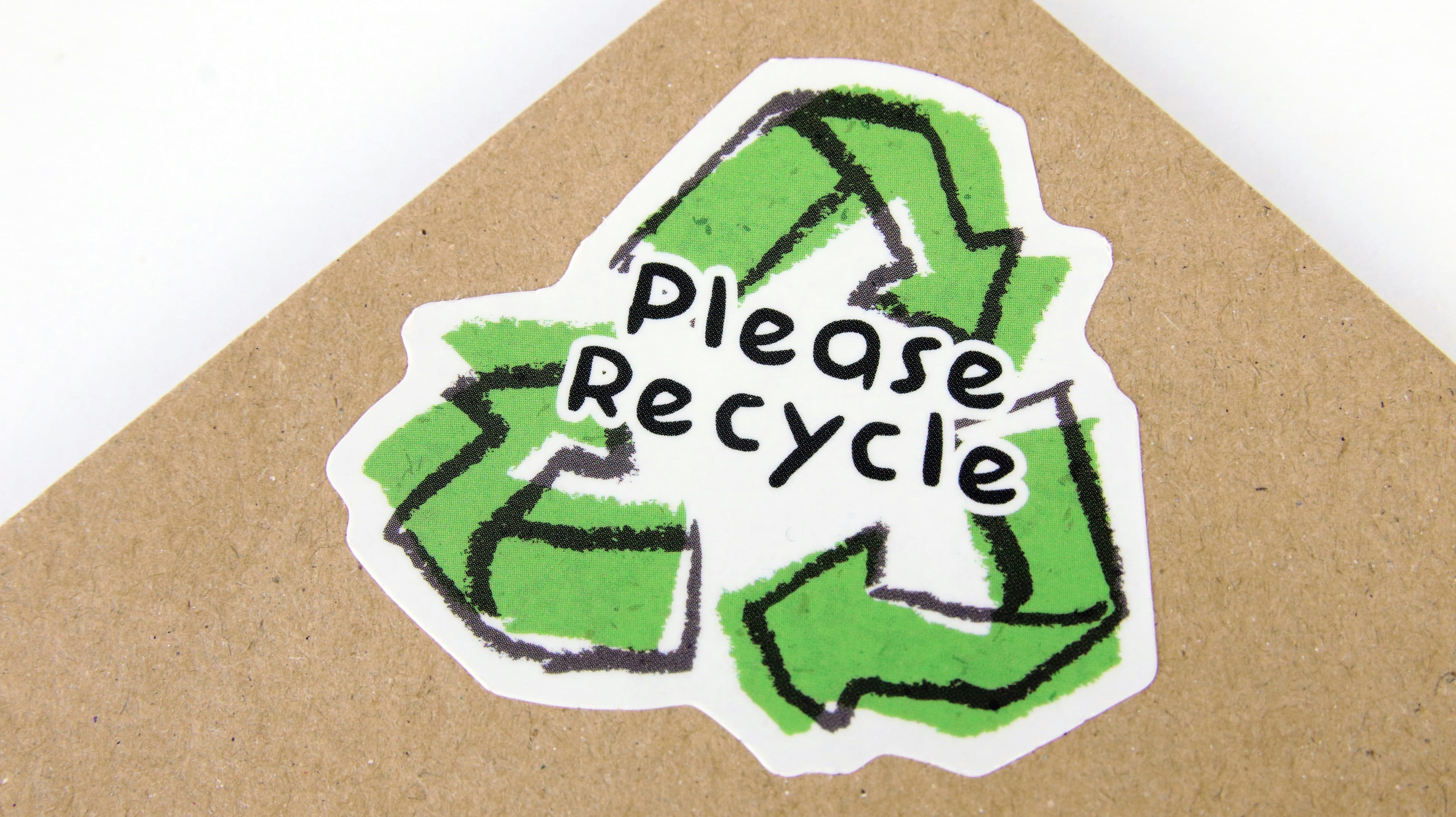 Auf einem Aufkleber auf braunem Karton ist ein grünes Recycling-Symbol zu sehen, über das in schwarzen, markiererstiligen Buchstaben die Aufschrift „Bitte recyceln“ geschrieben steht.