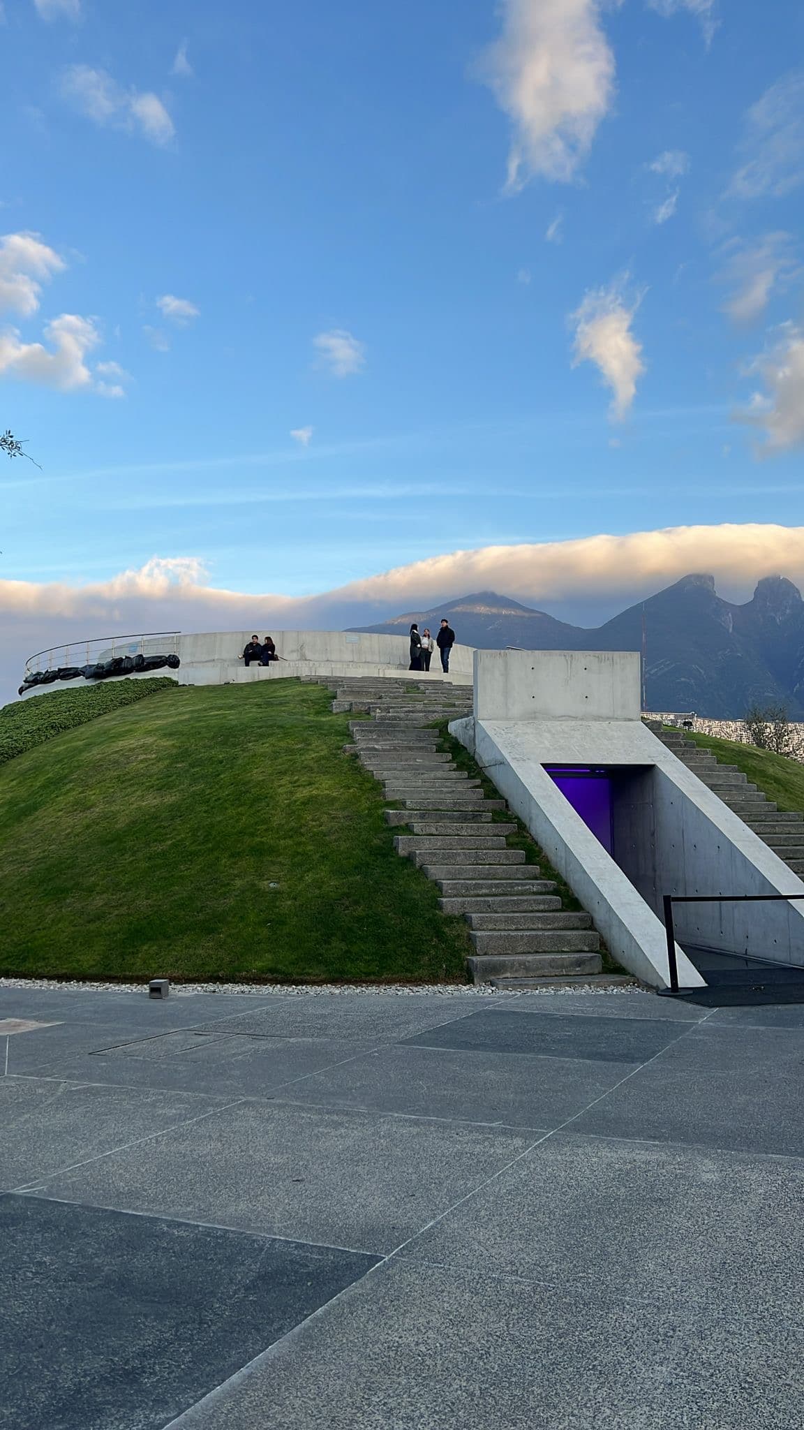 Ein grasbewachsener Hügel mit Betontreppen und einem modernen Bauwerk, dessen Eingang in violettes Licht getaucht ist; oben stehen Menschen, im Hintergrund sind Berge unter einem teilweise bewölkten Himmel zu sehen.