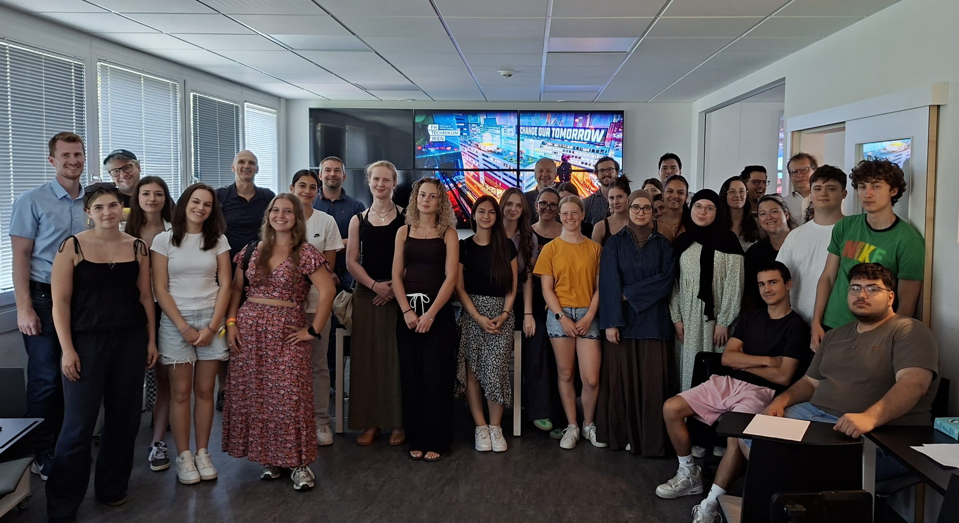 Eine Gruppe von Menschen posiert für ein Foto in einem Klassenzimmer, im Hintergrund ist ein großer Digitalbildschirm zu sehen, auf dem Grafiken angezeigt werden. Einige stehen, andere sitzen, und alle blicken lächelnd in die Kamera.