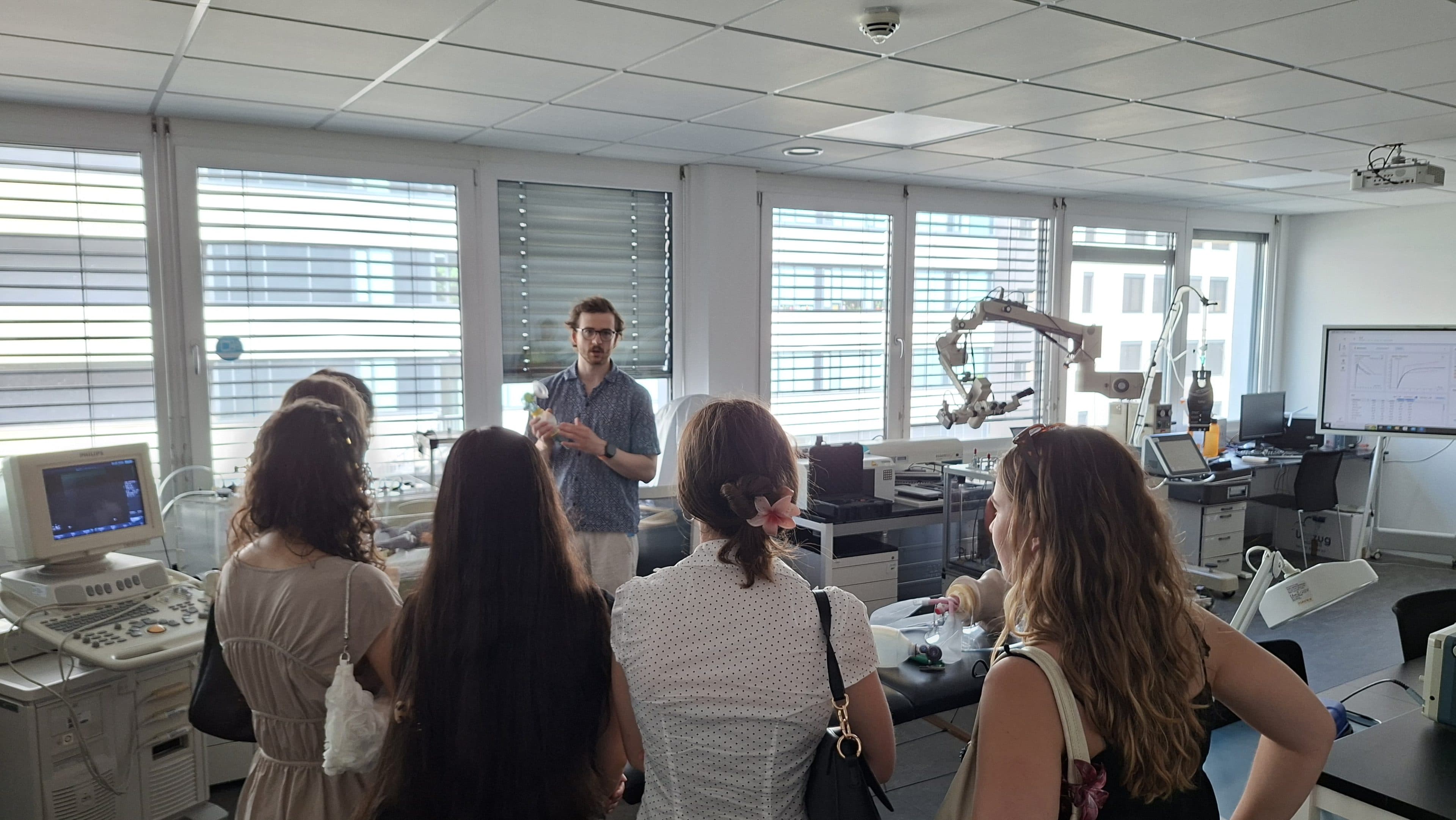 Ein Mann spricht zu einer Gruppe von Frauen in einem modernen Labor, das mit medizinischen Geräten ausgestattet ist und im Hintergrund große Fenster sowie Computermonitore aufweist.