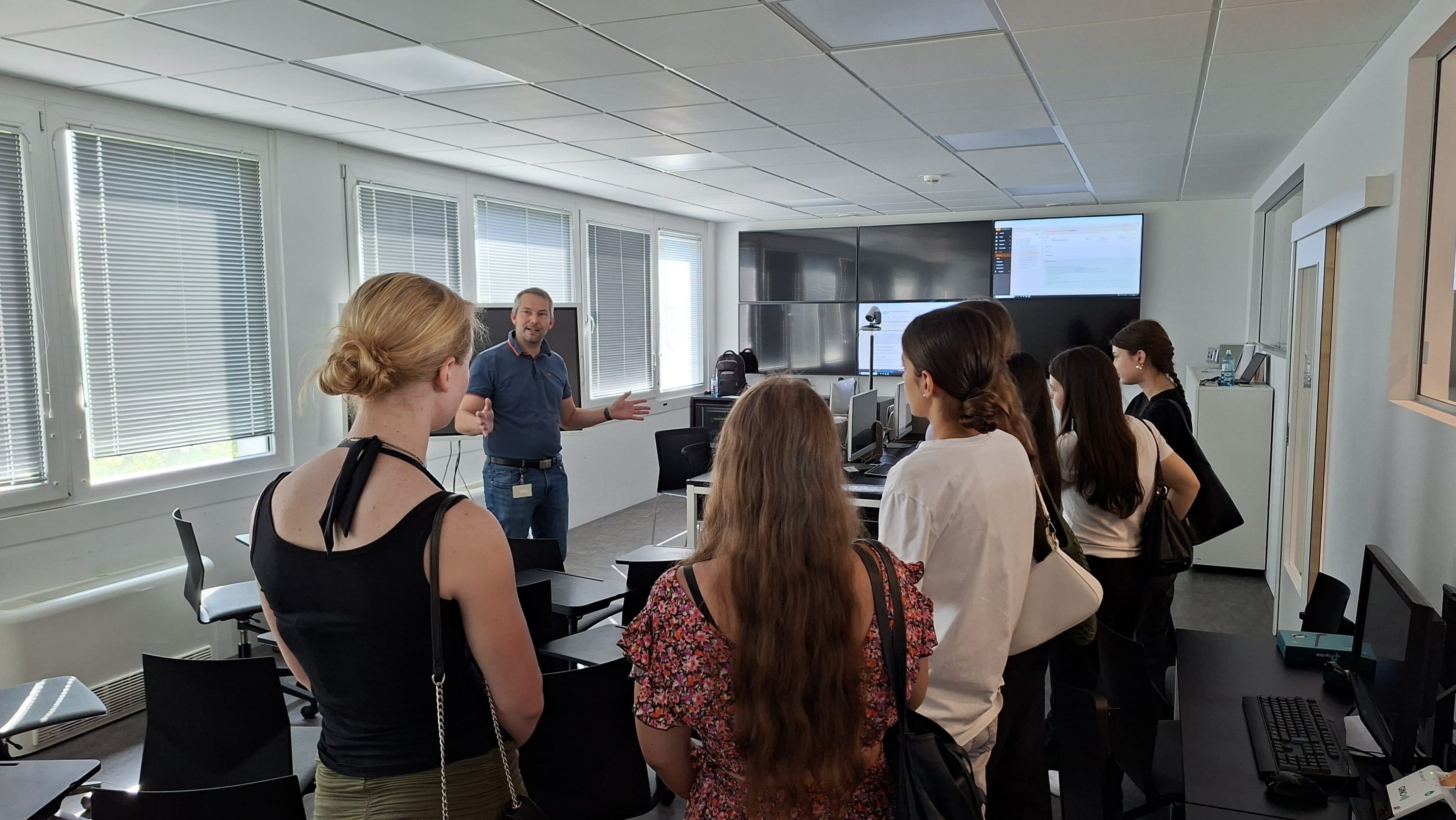 Ein Mann spricht zu einer Gruppe von fünf Frauen in einem modernen, hellen Büro-Schulungsraum mit Schreibtischen, Computern und großen Fenstern.