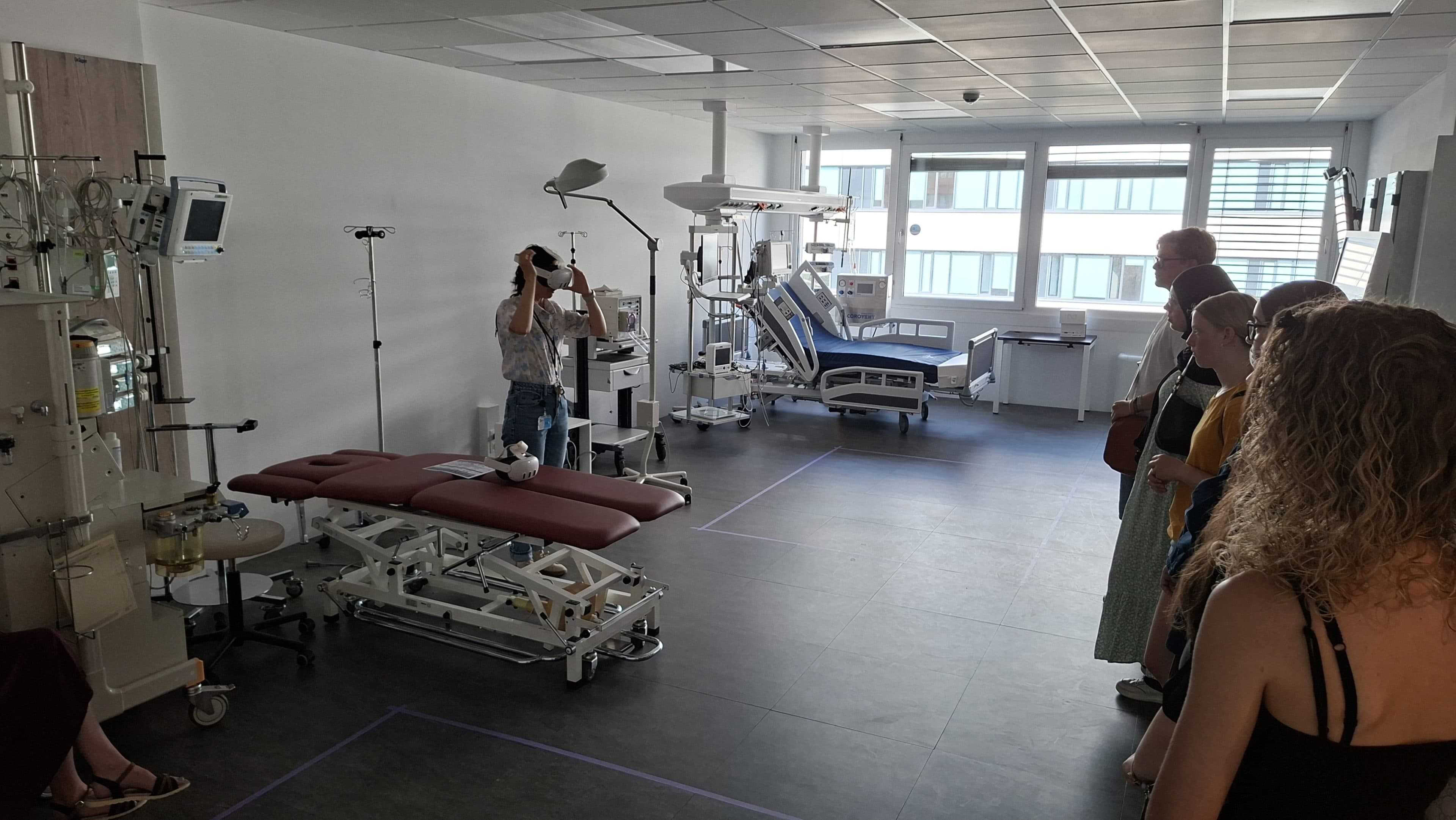 Eine Person führt in einem Krankenhauszimmer medizinische Geräte vor, während eine Gruppe von Menschen in einer Reihe steht und zusieht. Im Raum befinden sich Betten, Monitore und andere medizinische Geräte. Durch große Fenster fällt Sonnenlicht herein.