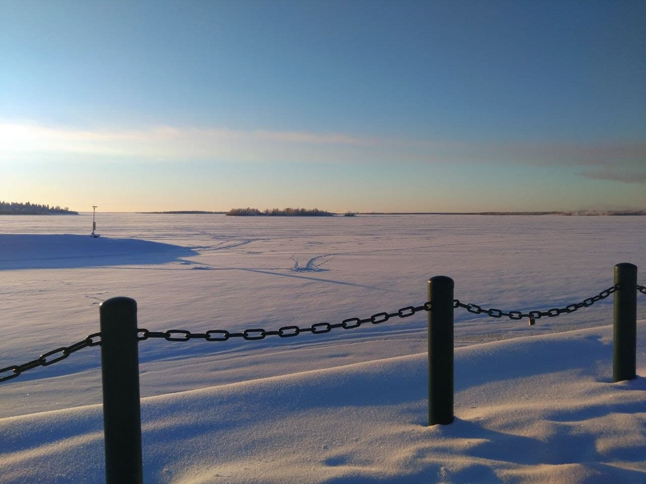 Unter einem strahlend blauen Himmel erstreckt sich eine verschneite, flache Landschaft, in der vereinzelt Bäume und leichte Spuren im Schnee zu sehen sind. Im Vordergrund verläuft eine schwarze Maschendrahtbarriere.