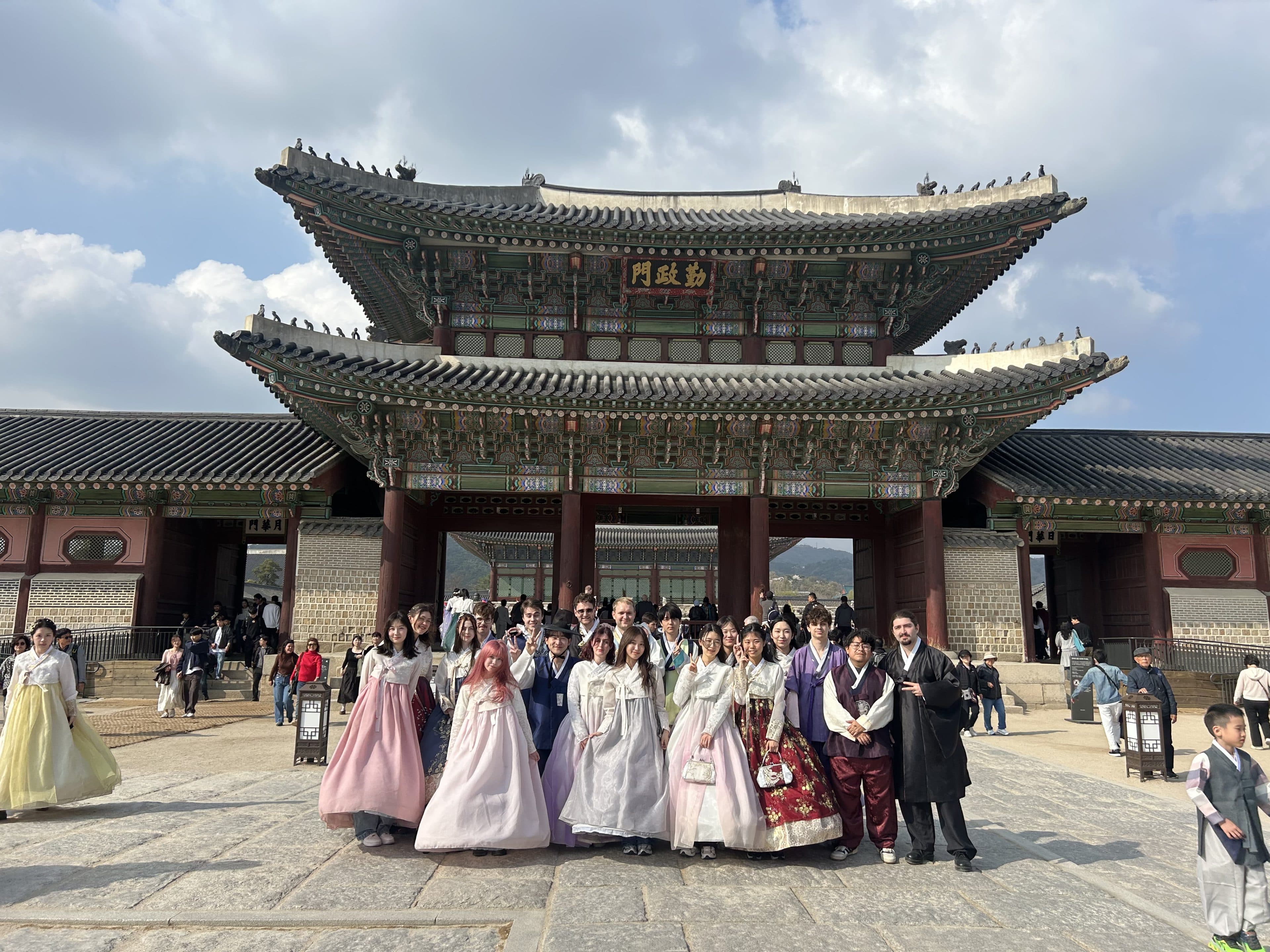 An einem sonnigen Tag posiert eine Gruppe von Menschen in traditioneller und moderner Kleidung vor einem großen, reich verzierten koreanischen Palasttor.