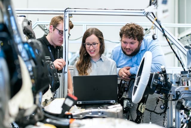 Drei Personen blicken auf einen Laptop, während sie in einem Innenraum an einer Maschine arbeiten. Sie scheinen Daten zu besprechen oder zu analysieren, die mit der Maschine zusammenhängen, deren verschiedene Bauteile freiliegen.