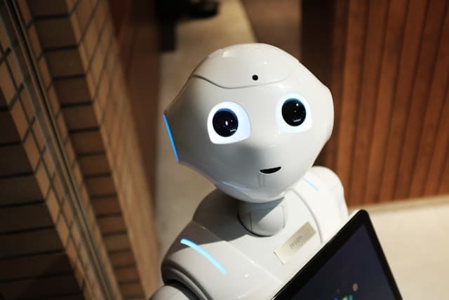 Ein weißer humanoider Roboter mit leuchtend blauen Augen blickt mit einem Tablet in der Hand nach oben. Er steht in einem Innenraum neben einer Backsteinwand und Holzvertäfelungen.