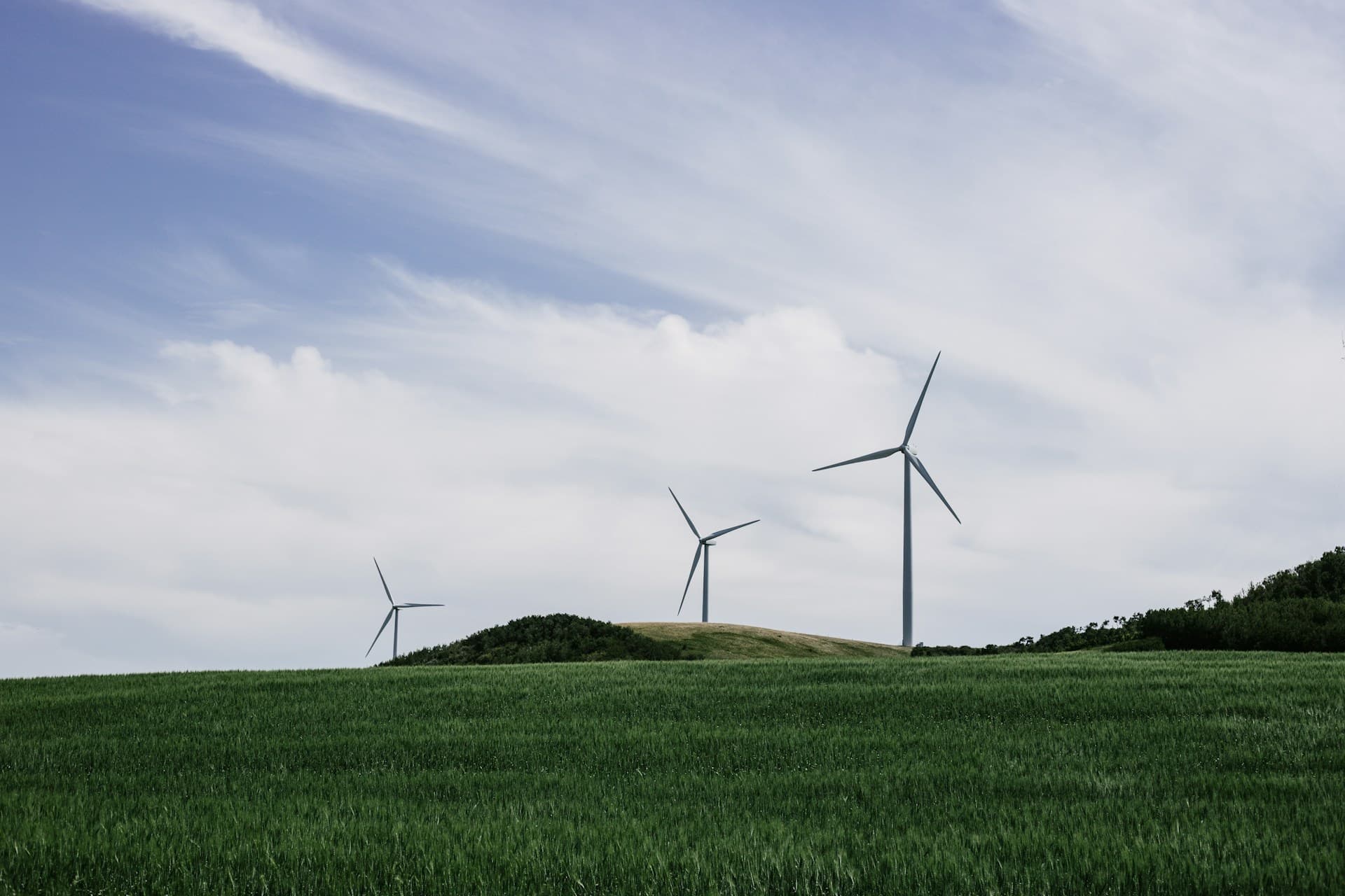 Drei große Windkraftanlagen stehen auf grasbewachsenen Hügeln unter einem teilweise bewölkten Himmel, umgeben von grünen Feldern und sanften Hängen.