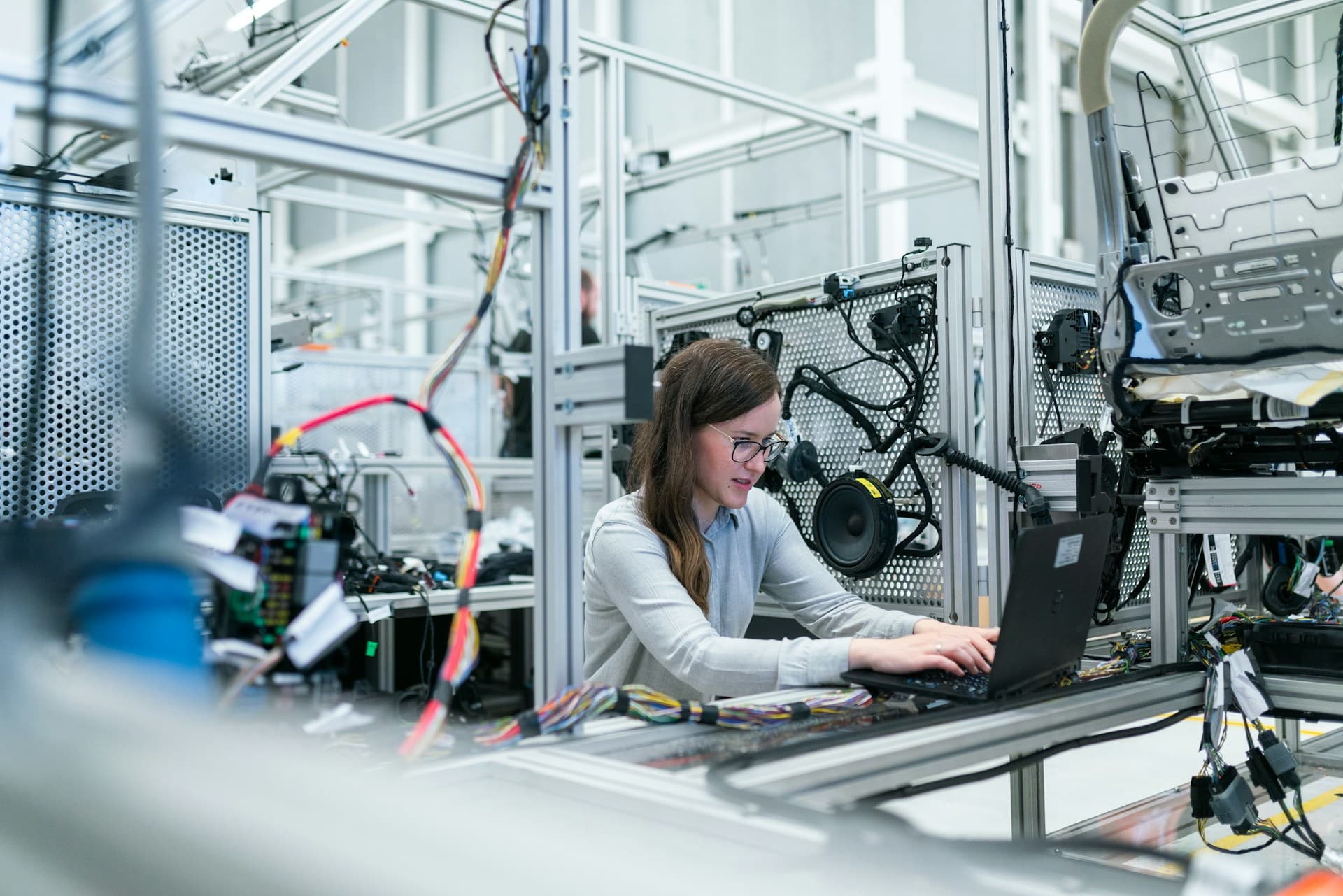 Eine Frau arbeitet an einem Laptop, umgeben von Maschinen und Kabeln in einer modernen Industrie- oder Laborumgebung, was auf ingenieurwissenschaftliche oder technische Forschungsarbeit hindeutet.