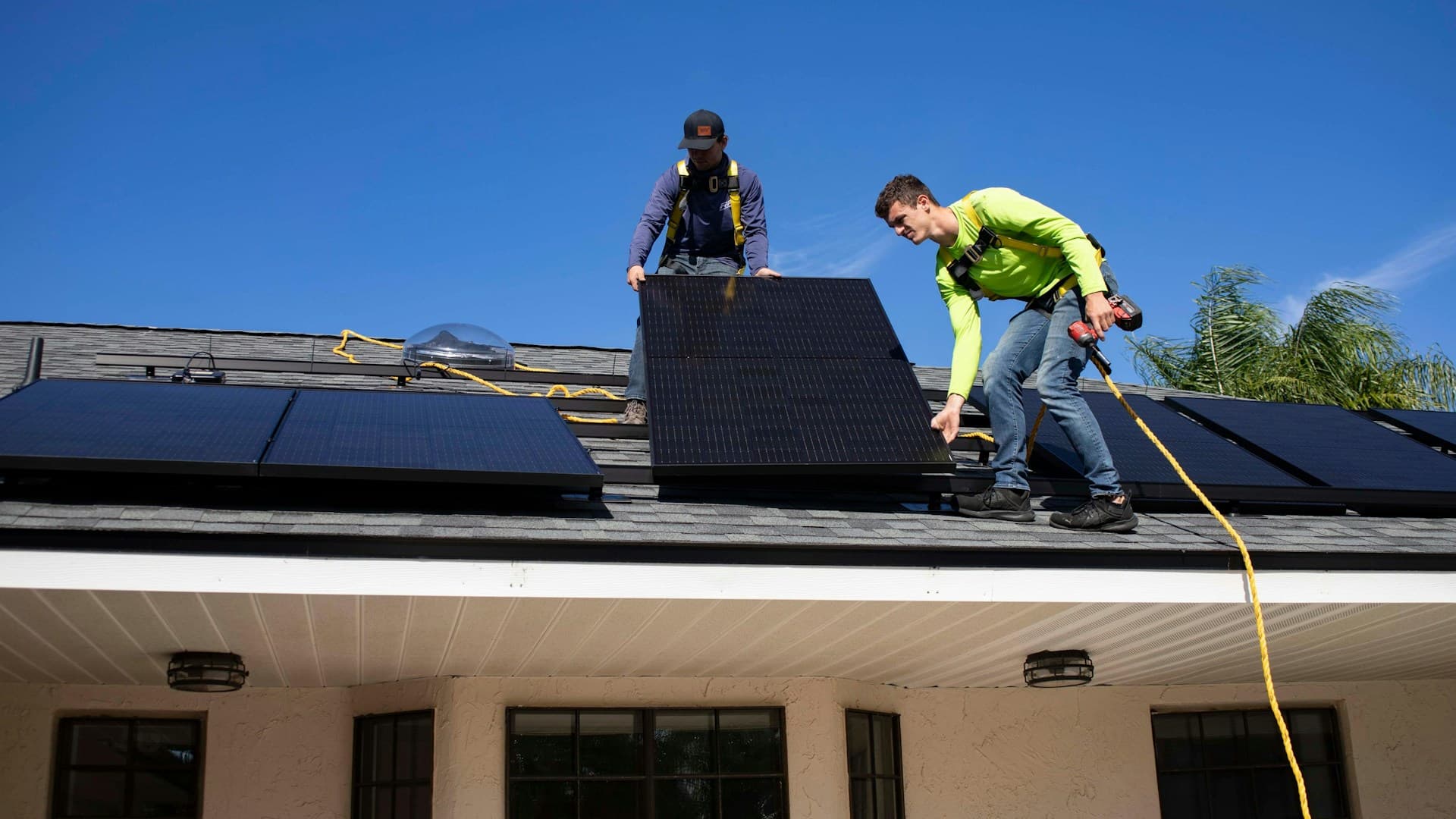 Unter strahlend blauem Himmel montieren zwei Arbeiter mit Sicherheitsgurten und Werkzeugen Solarmodule auf einem Hausdach.