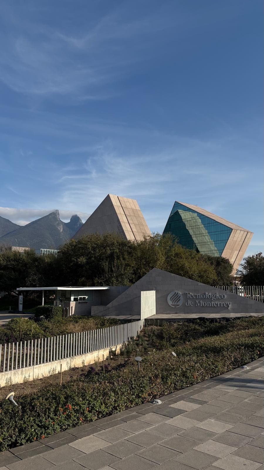 Moderne Glas- und Betongebäude des Tecnológico de Monterrey mit Bergen im Hintergrund, Bäumen und Sträuchern im Mittelgrund und einem gepflasterten Gehweg im Vordergrund unter blauem Himmel.
