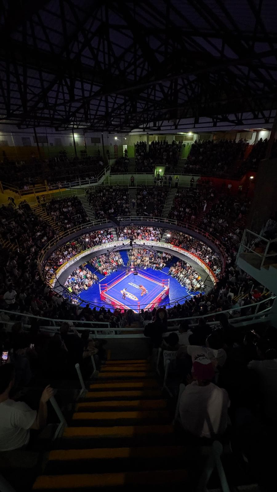 Eine große Menschenmenge umringt einen hell beleuchteten Boxring in der Mitte einer Halle, gesehen von einem hohen Aussichtspunkt auf der Tribüne.