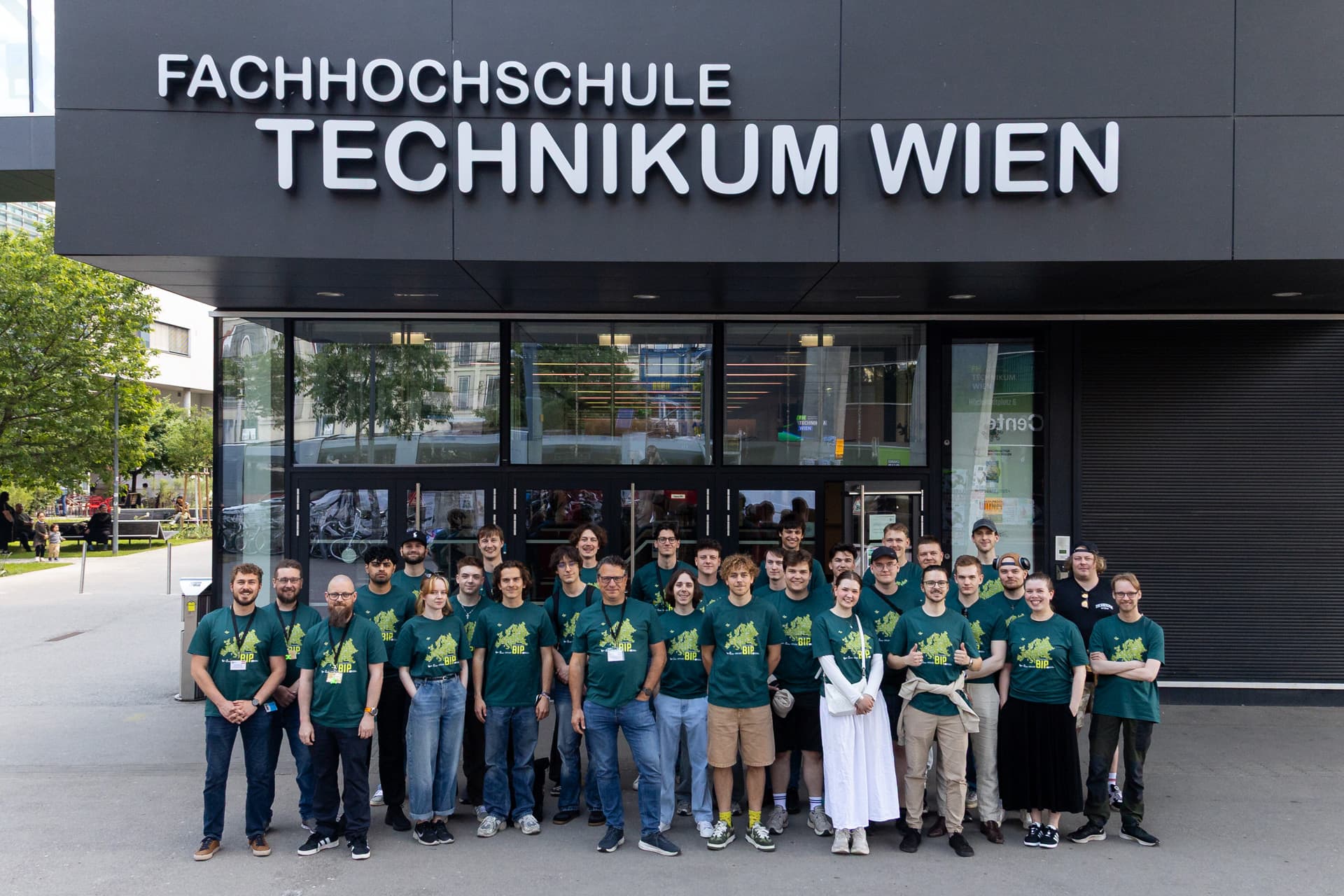 Eine Gruppe von Menschen in einheitlichen grünen T-Shirts steht lächelnd vor dem Eingang des Gebäudes der Fachhochschule Technikum Wien.
