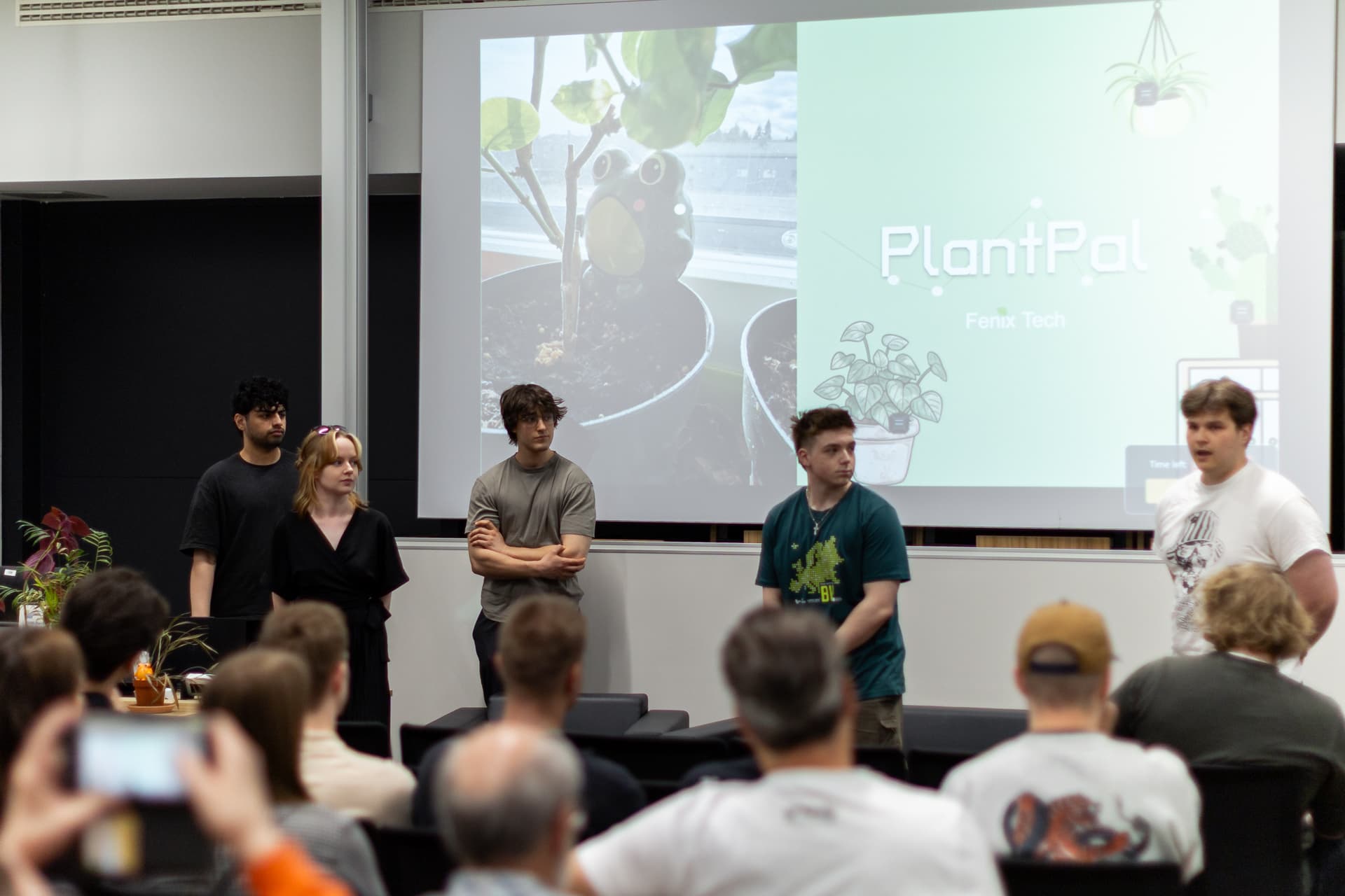 Fünf Personen stehen vor einem Publikum und stellen ein Projekt namens „PlantPal“ vor, während hinter ihnen auf der Leinwand eine Folie mit Pflanzenmotiven zu sehen ist. Einige Zuschauer machen Fotos oder hören aufmerksam zu.