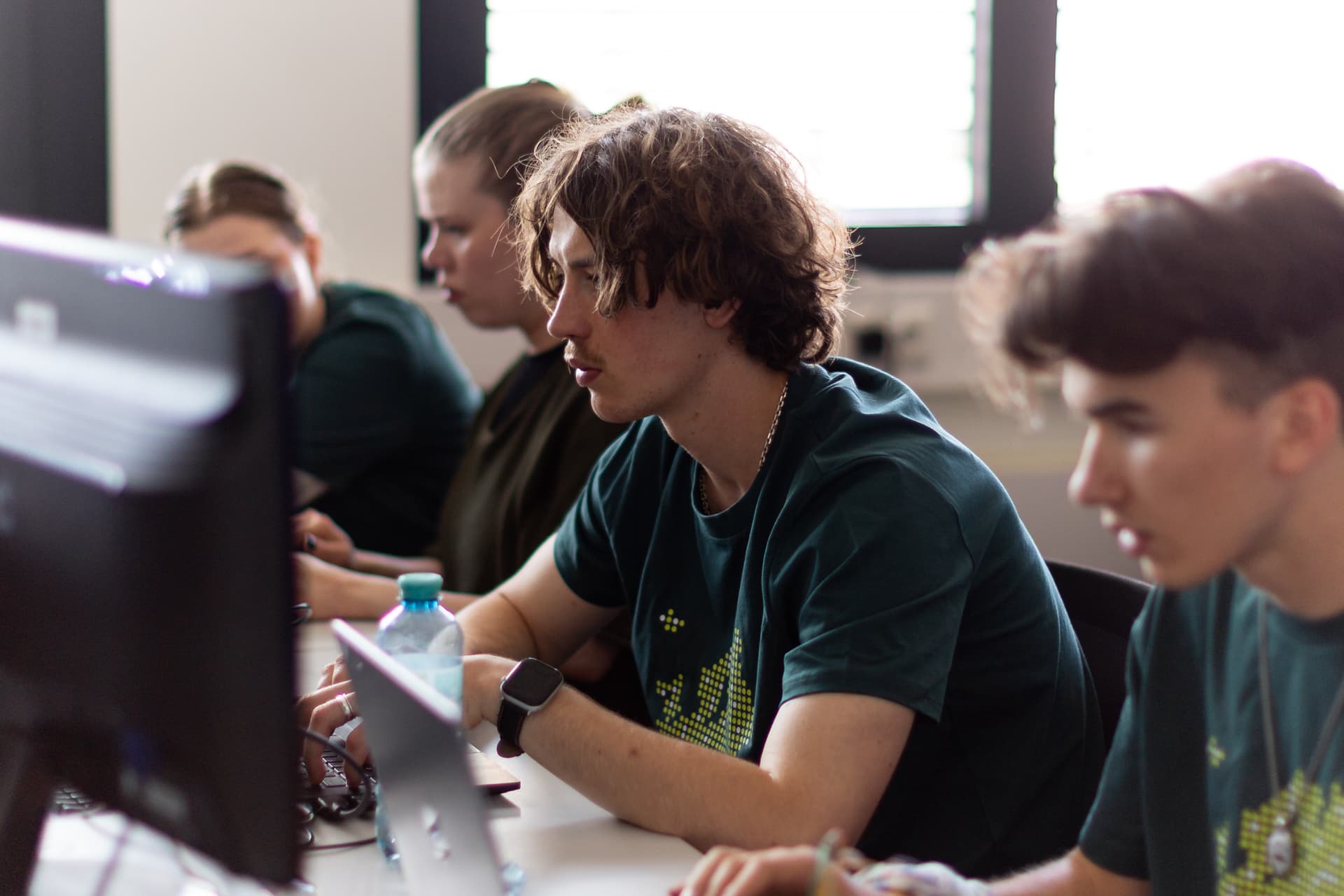 Vier junge Menschen sitzen an einem Schreibtisch und blicken konzentriert auf ihre Computerbildschirme. Sie tragen dunkelgrüne Hemden und arbeiten gemeinsam in einem hell beleuchteten Raum mit großen Fenstern, in dem moderne Geräte zu sehen sind.