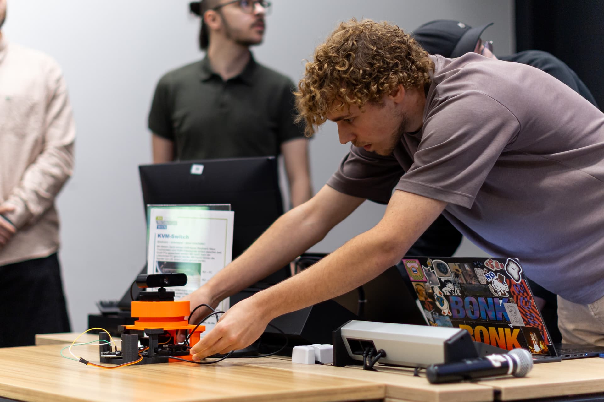 Eine Person stellt ein orangefarbenes Gerät mit Kabeln auf einem Tisch ein, neben einem Laptop, der mit Aufklebern, einem Schild und einem Mikrofon bedeckt ist. Im Hintergrund sind weitere Personen und Bildschirme zu sehen.