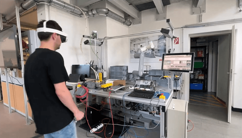Eine Person, die eine Augmented-Reality-Brille trägt, steht vor einem Elektronik-Arbeitsplatz mit verschiedenen Kabeln, Monitoren und Geräten in einer modernen Industrielaborumgebung.