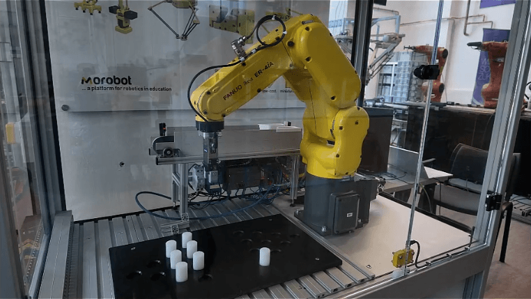 Ein gelber Roboterarm legt weiße, zylindrische Gegenstände auf ein schwarzes Tablett in einem Glasgehäuse in einem Labor oder einer Bildungseinrichtung.