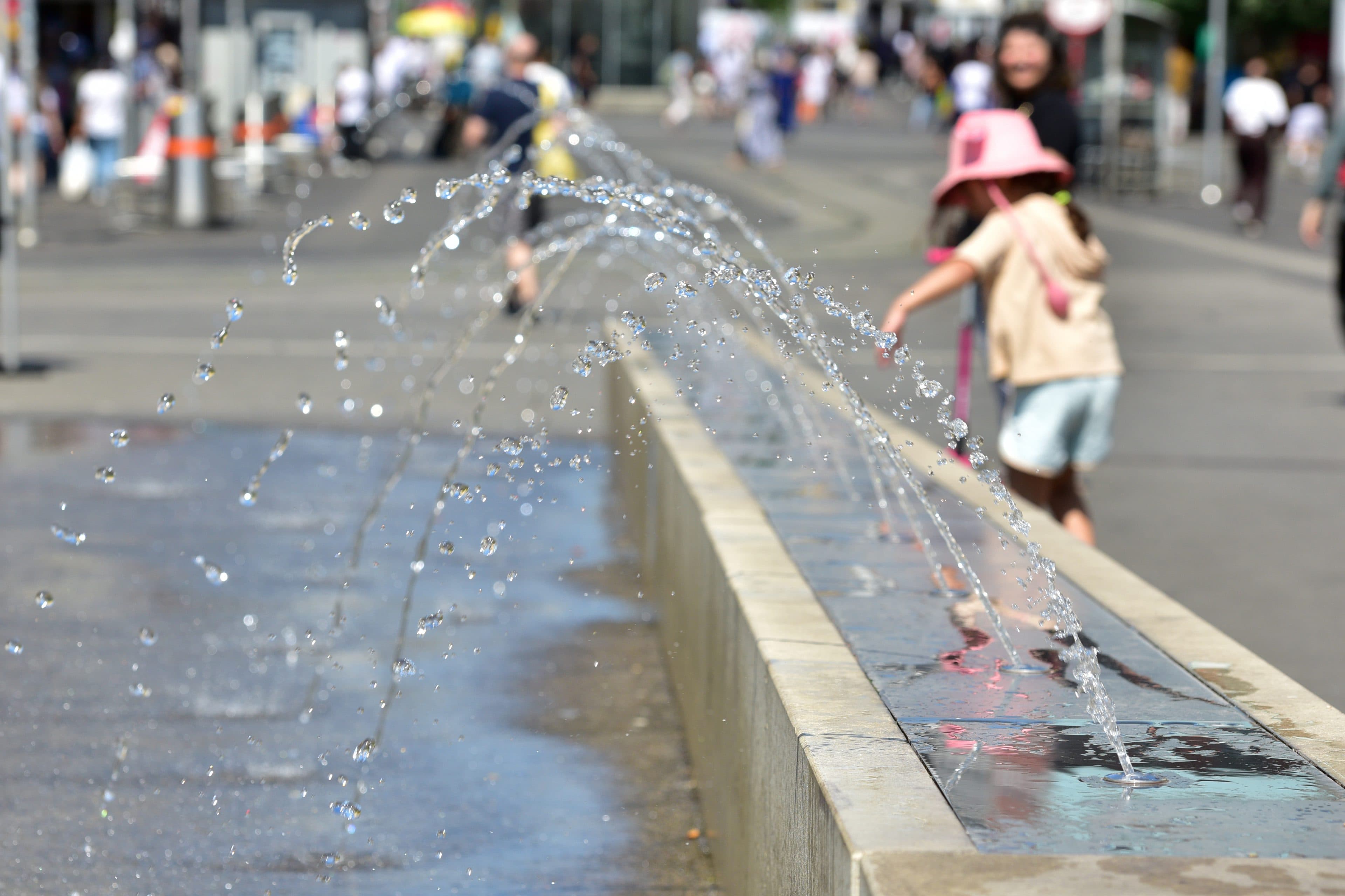 Zwei Kinder in Sommerkleidung spielen an einem sonnigen Tag in der Nähe einer Reihe von Wasserstrahlen in einem öffentlichen Springbrunnen im Freien, während im Hintergrund Menschen und Gebäude verschwommen zu sehen sind.