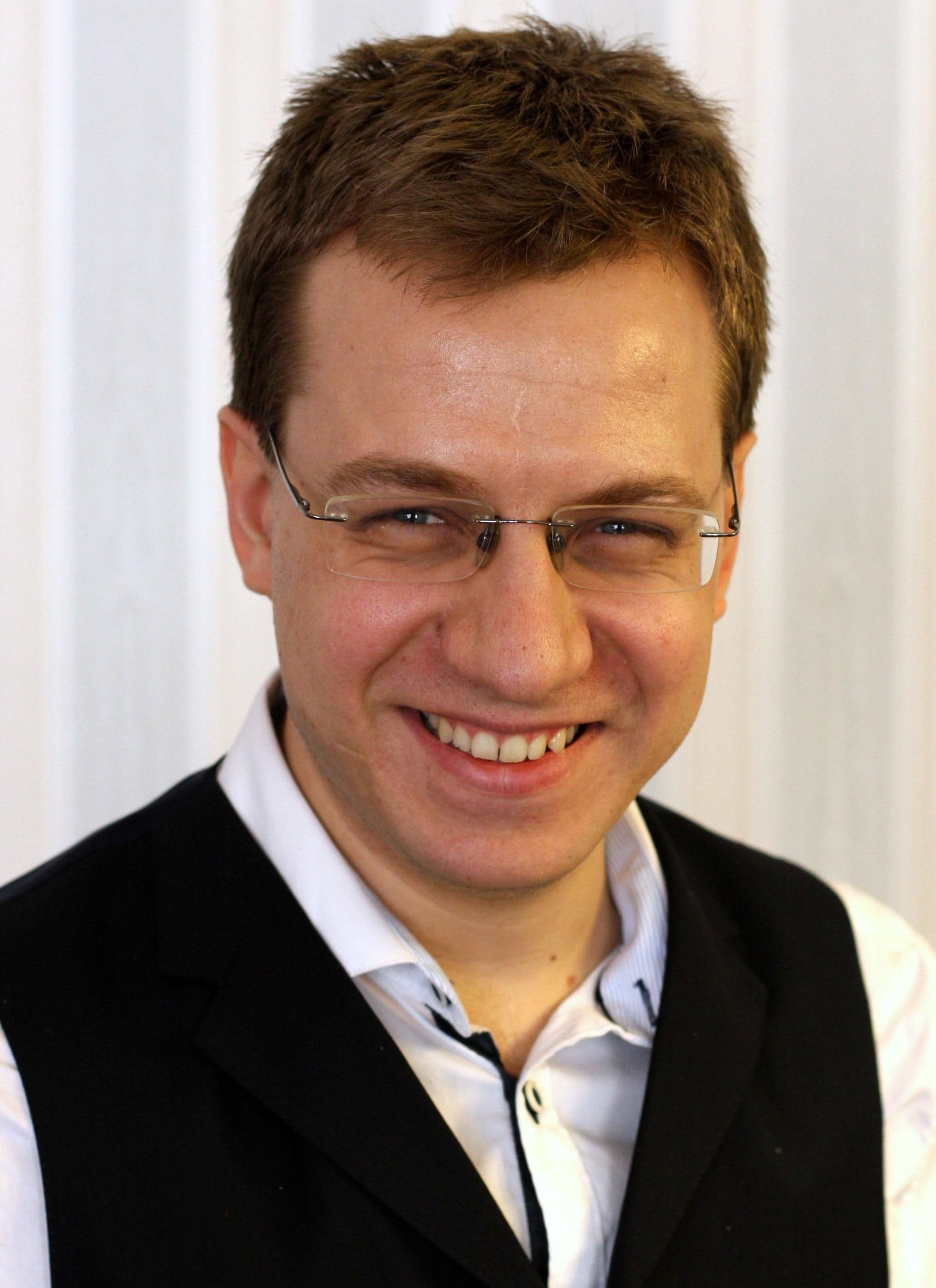 Ein Mann mit kurzen braunen Haaren, Brille und einem weißen Hemd unter einer schwarzen Weste lächelt in die Kamera. Im Hintergrund ist eine helle, vertikal gestreifte Wand zu sehen.