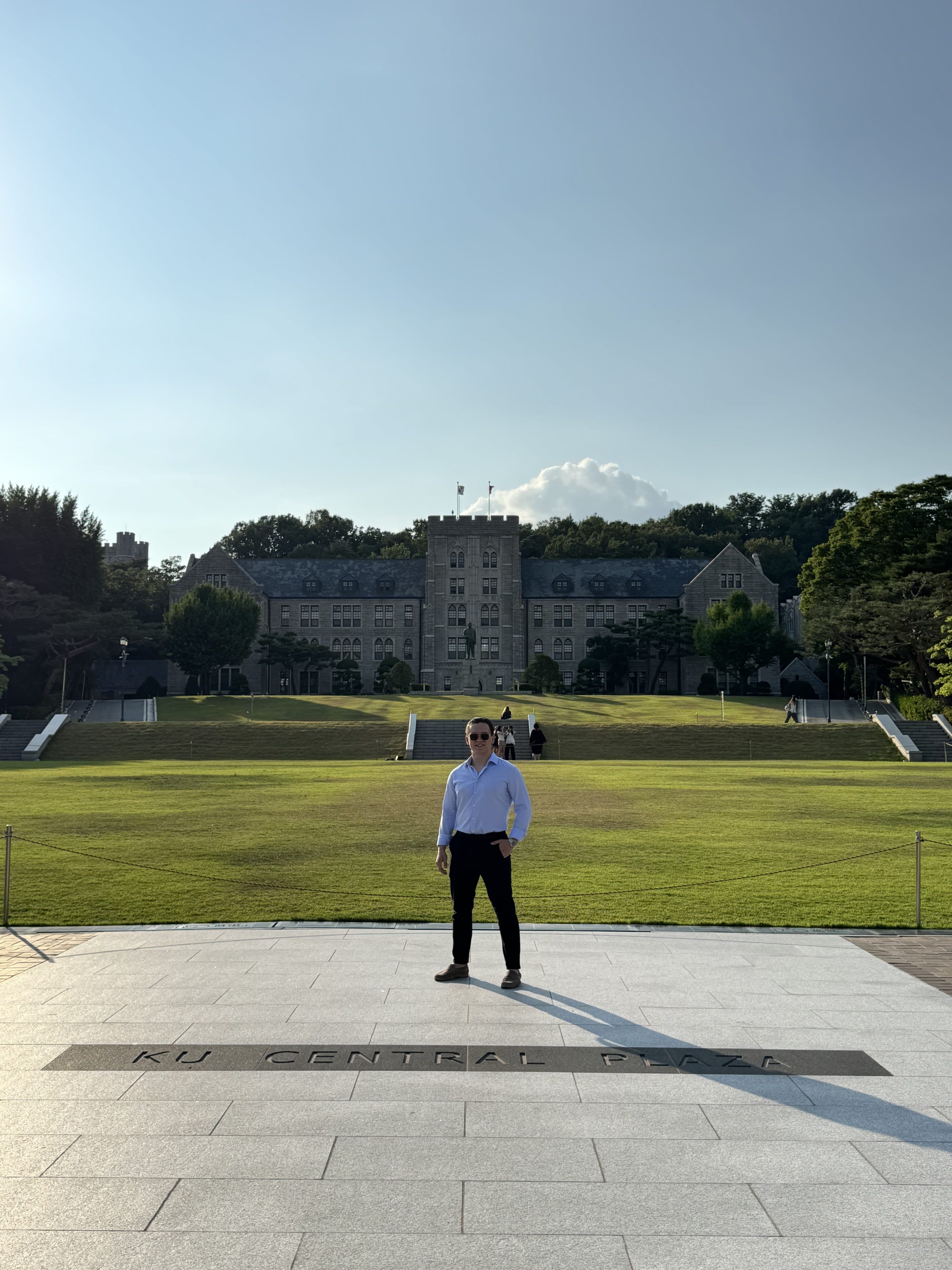 Eine Person mit Sonnenbrille steht auf einem Platz mit der Aufschrift „KU CENTRAL PLAZA“, im Hintergrund sind unter strahlend blauem Himmel ein großes, historisches Steingebäude und eine grüne Rasenfläche zu sehen.