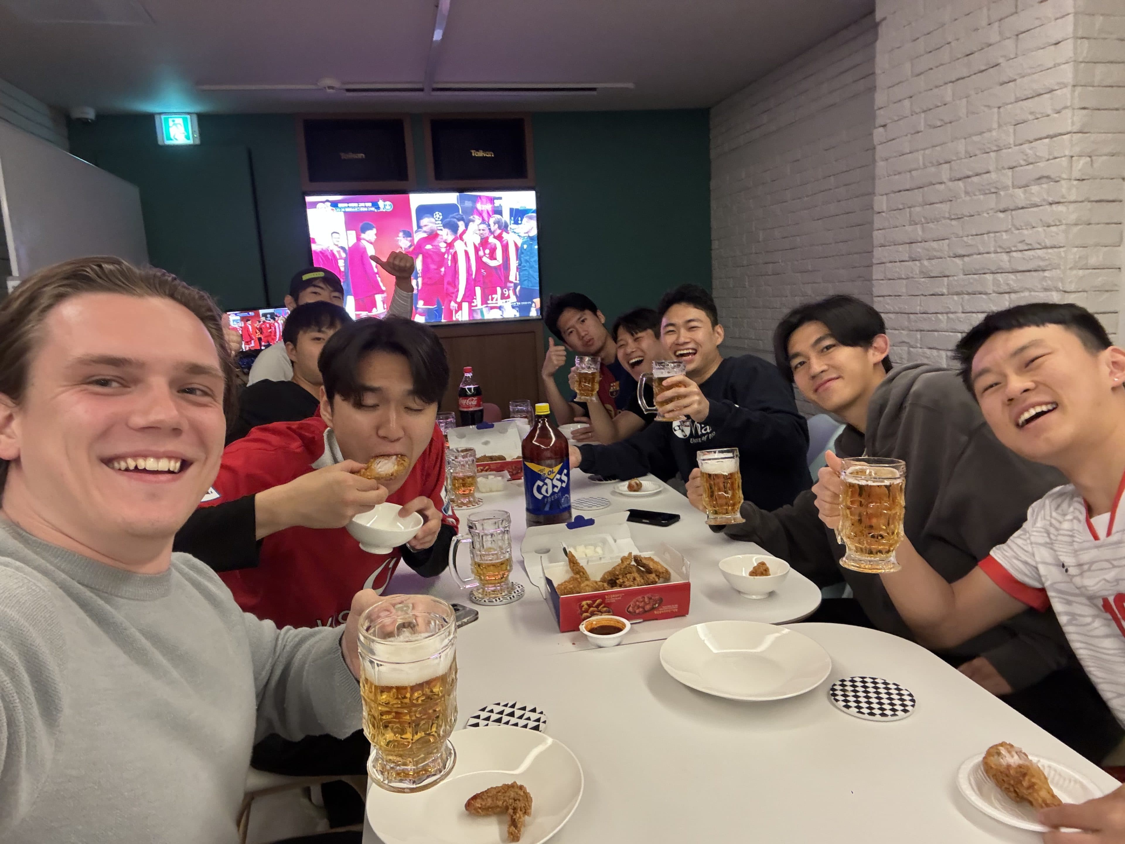 Eine Gruppe von Freunden sitzt mit Bier, Brathähnchen und Snacks um einen Tisch herum, lächelt und stößt mit den Gläsern an. Im Hintergrund läuft im Fernsehen ein Fußballspiel.