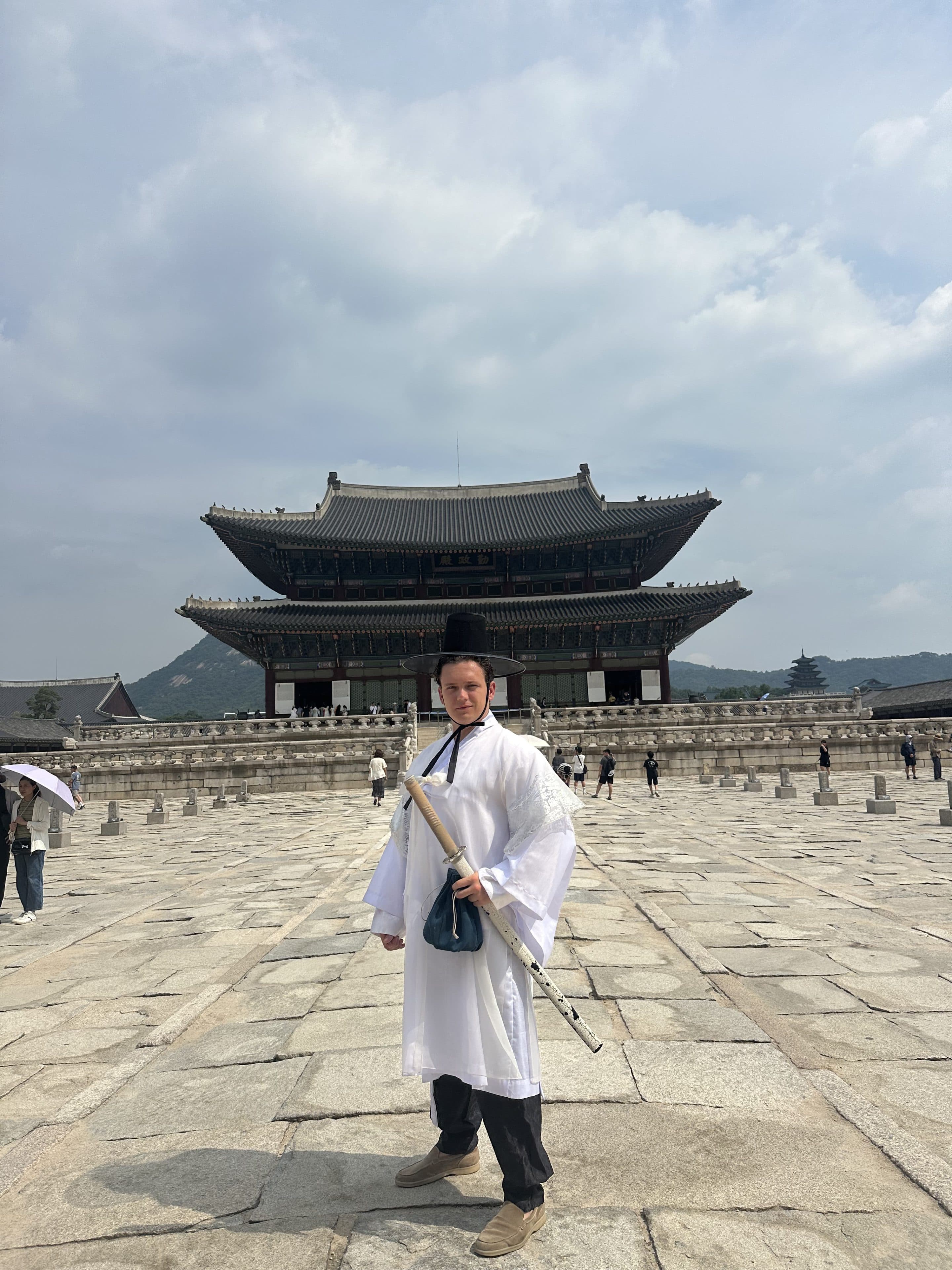 Eine Person in traditioneller koreanischer Tracht posiert mit einem Schwert vor dem Gyeongbokgung-Palast in Seoul, Südkorea. Im Hintergrund sind unter einem bewölkten Himmel weitere Besucher und Berge zu sehen.