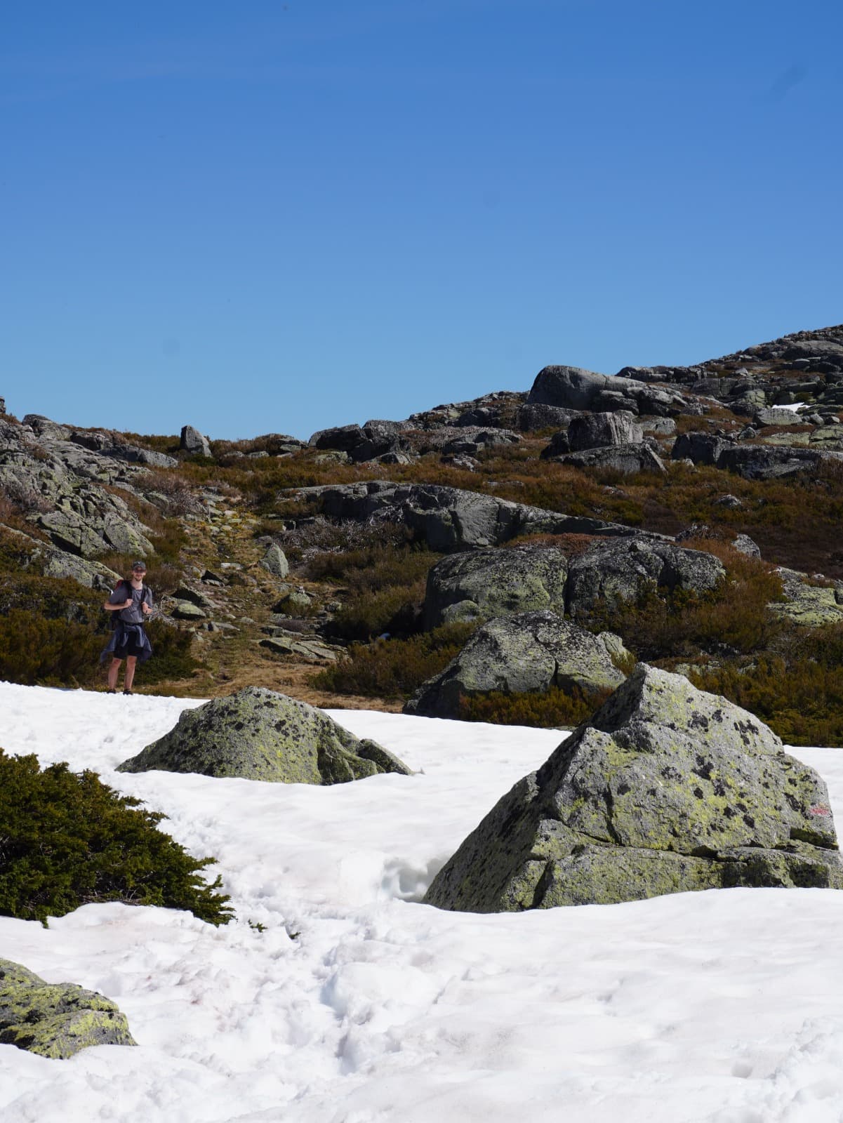 Eine Person steht in einer felsigen Berglandschaft, umgeben von Schneeflecken und großen Felsbrocken unter einem strahlend blauen Himmel.
