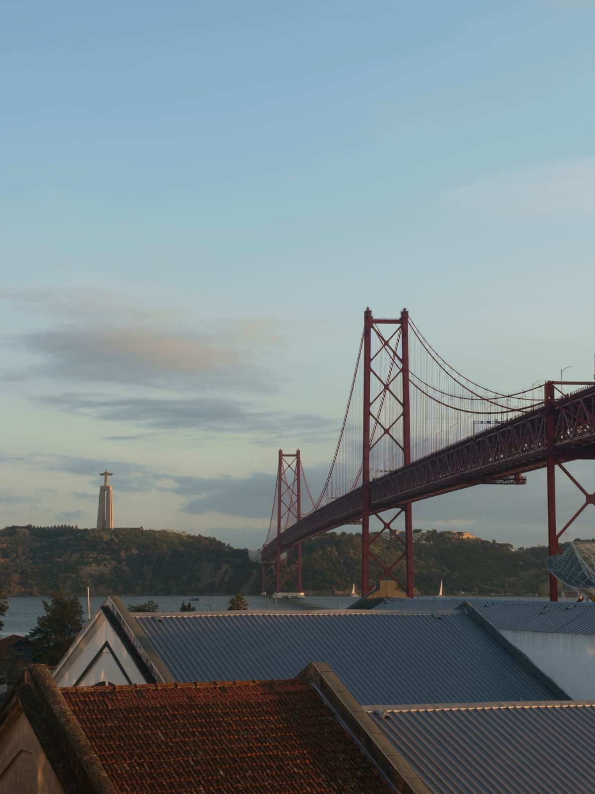 Eine rote Hängebrücke spannt sich bei Sonnenuntergang über das Wasser, im Vordergrund sind Hausdächer zu sehen. Auf einem entfernten Hügel steht unter einem teilweise bewölkten Himmel eine große Christusstatue.
