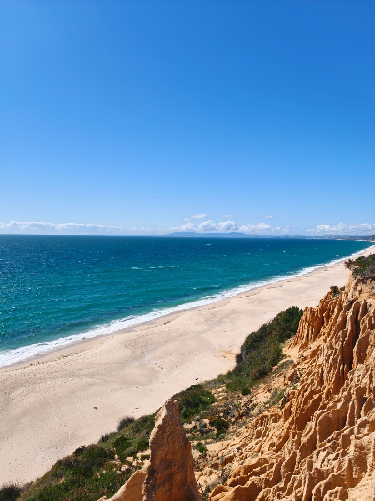 Blick von sandigen Klippen auf einen weiten, menschenleeren Strand mit türkisfarbenen Meereswellen unter einem strahlend blauen Himmel mit vereinzelten Wolken.
