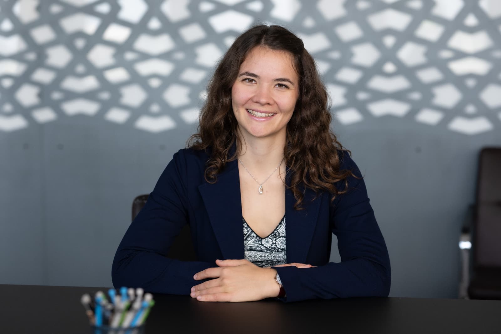 Eine Frau mit langen Locken, die einen marineblauen Blazer trägt, sitzt lächelnd an einem Schreibtisch. Im Hintergrund ist ein geometrisches Lichtmuster an der Wand zu sehen. Im Vordergrund sind Stifte zu erkennen.
