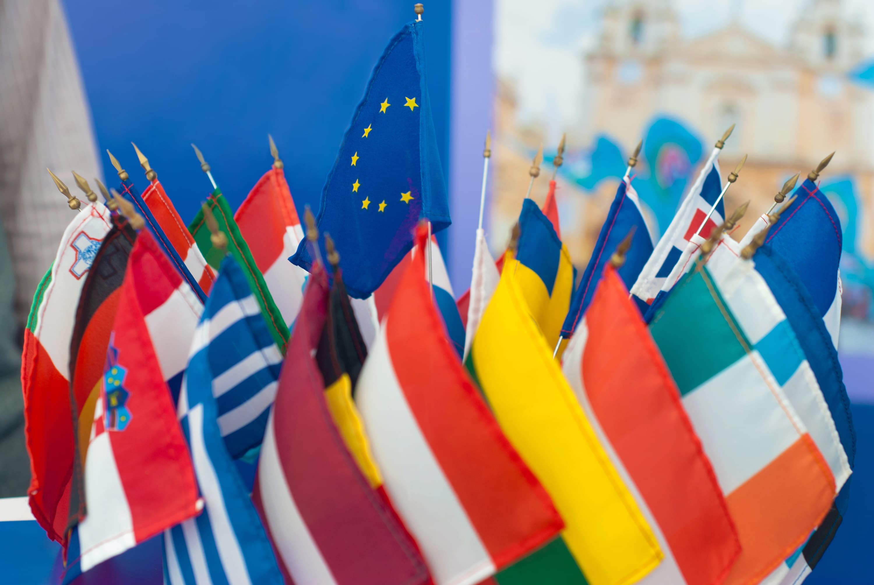 Mehrere kleine internationale Flaggen, darunter die Flaggen europäischer Länder und der Europäischen Union, sind dicht beieinander an Fahnenmasten zu sehen, vor einem verschwommenen blau-weißen Hintergrund.