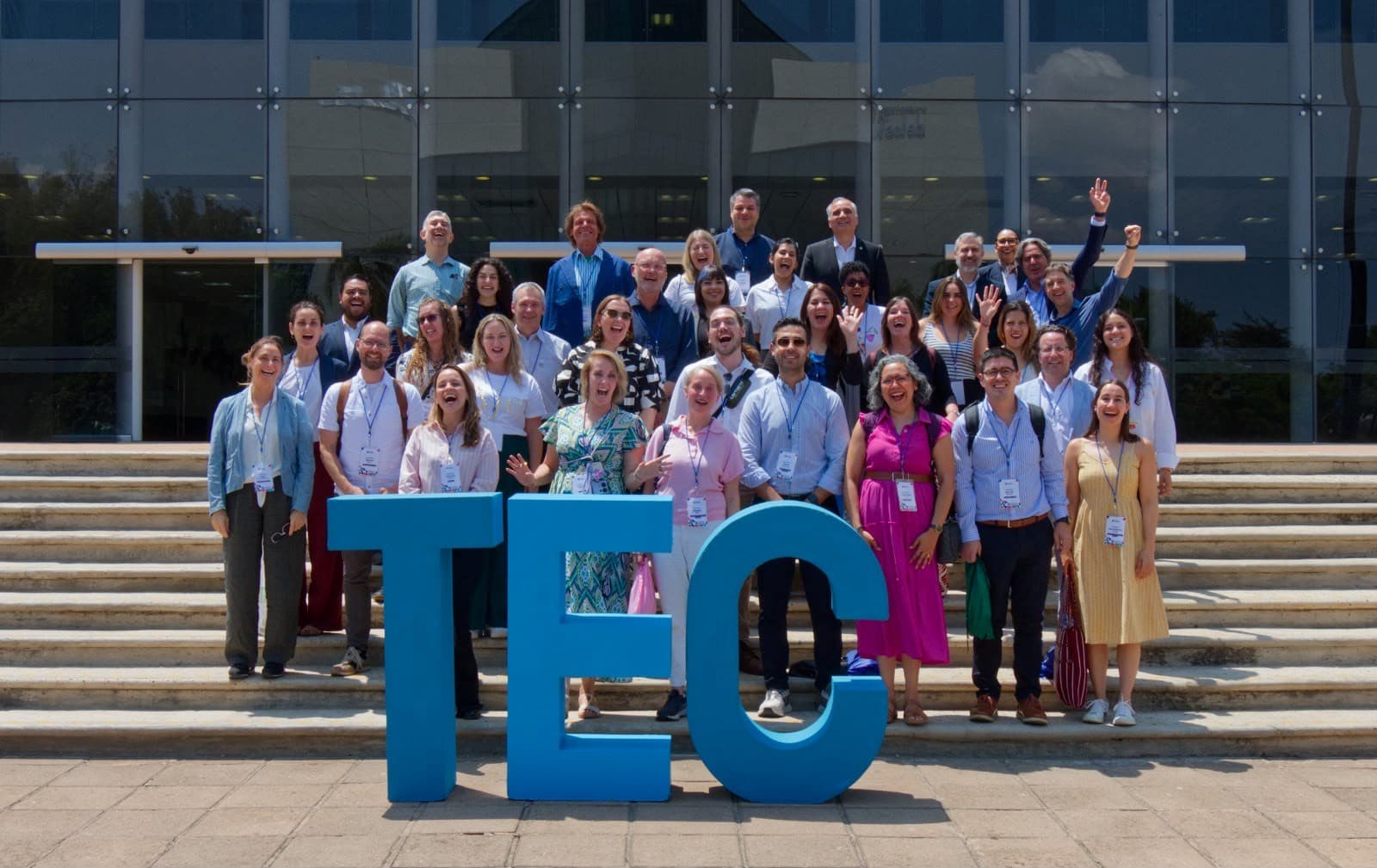 Eine große Gruppe von Menschen steht lächelnd auf den Stufen vor einem Glasgebäude, vor dem drei große blaue Buchstaben die Abkürzung „TEC“ bilden. Viele tragen Namensschilder und Business-Casual-Kleidung und winken oder posieren fröhlich.