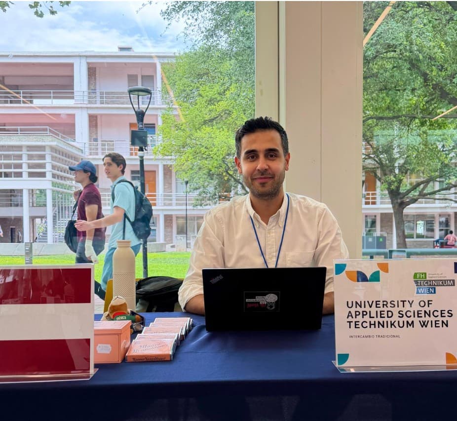 Ein Mann sitzt mit einem Laptop an einem Tisch neben einem Schild mit der Aufschrift „University of Applied Sciences Technikum Wien“ vor großen Fenstern, durch die man Menschen sehen kann, die draußen vorbeigehen.