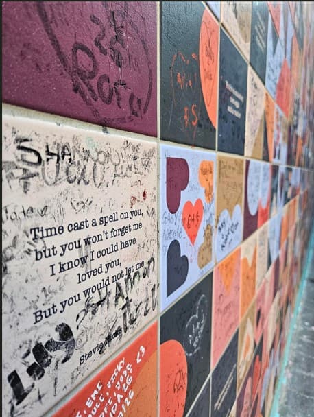 Eine bunte, mit Fliesen verkleidete Wand mit handgeschriebenen Botschaften und aufgedruckten Liebeszitaten, darunter: „Die Zeit hat dich verzaubert, aber du wirst mich nicht vergessen.“ „Ich weiß, ich hätte dich lieben können. Aber du hast es mir nicht erlaubt…“.