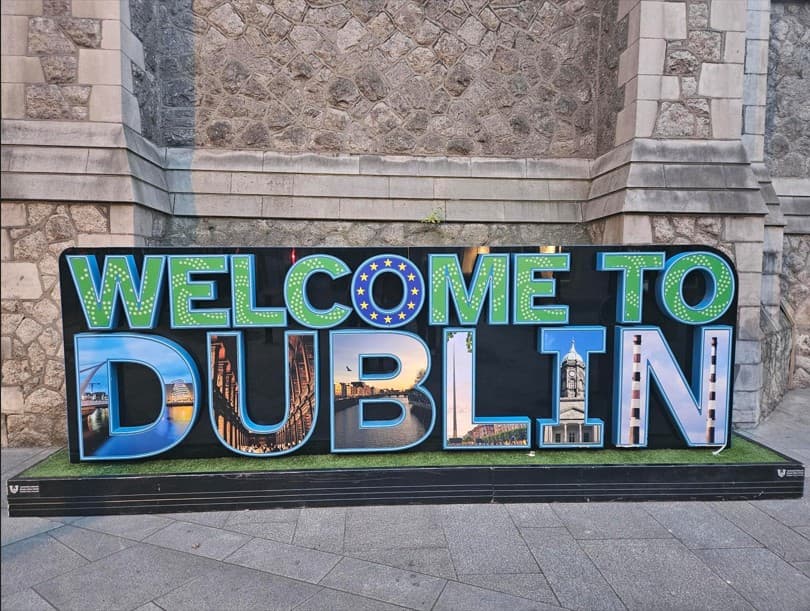Großes Schild mit der Aufschrift „Welcome to Dublin“, auf dem in den Buchstaben die Wahrzeichen der Stadt abgebildet sind, vor dem Hintergrund einer Steinmauer.