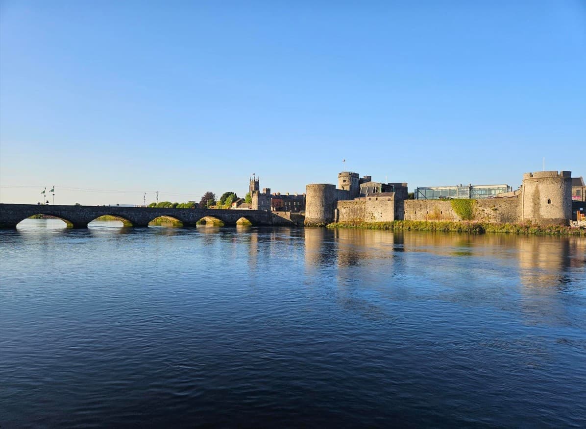 Eine Steinbrücke überspannt einen ruhigen Fluss und führt unter strahlend blauem Himmel zu einer historischen Steinburg mit Rundtürmen am gegenüberliegenden Ufer.