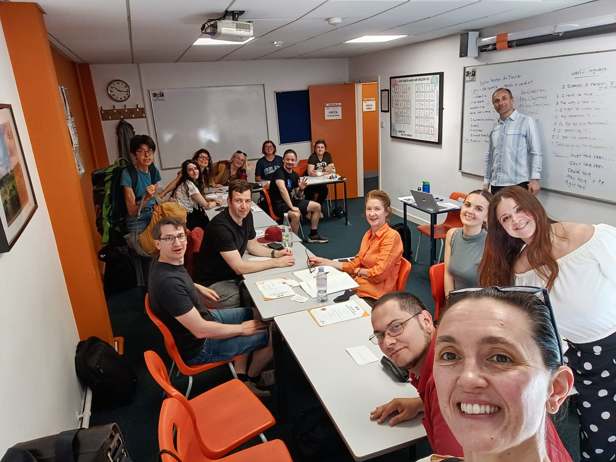 Eine Gruppe von Menschen lächelt und posiert für ein Selfie in einem Klassenzimmer mit orangefarbenen Stühlen, Whiteboards und Lehrplakaten. Einige sitzen an ihren Schreibtischen, andere stehen und beugen sich vor; sie wirken fröhlich und engagiert.