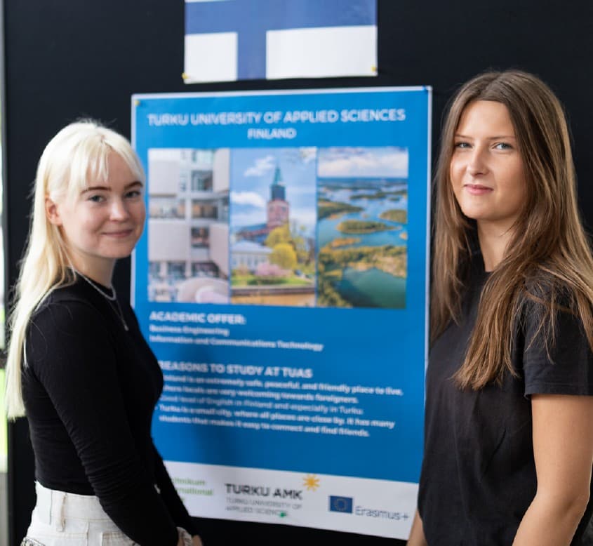 Zwei junge Frauen stehen vor einem Plakat der Fachhochschule Turku in Finnland und lächeln in die Kamera. Das Plakat zeigt Bilder vom Campus und listet das Studienangebot sowie Gründe für ein Studium dort auf.