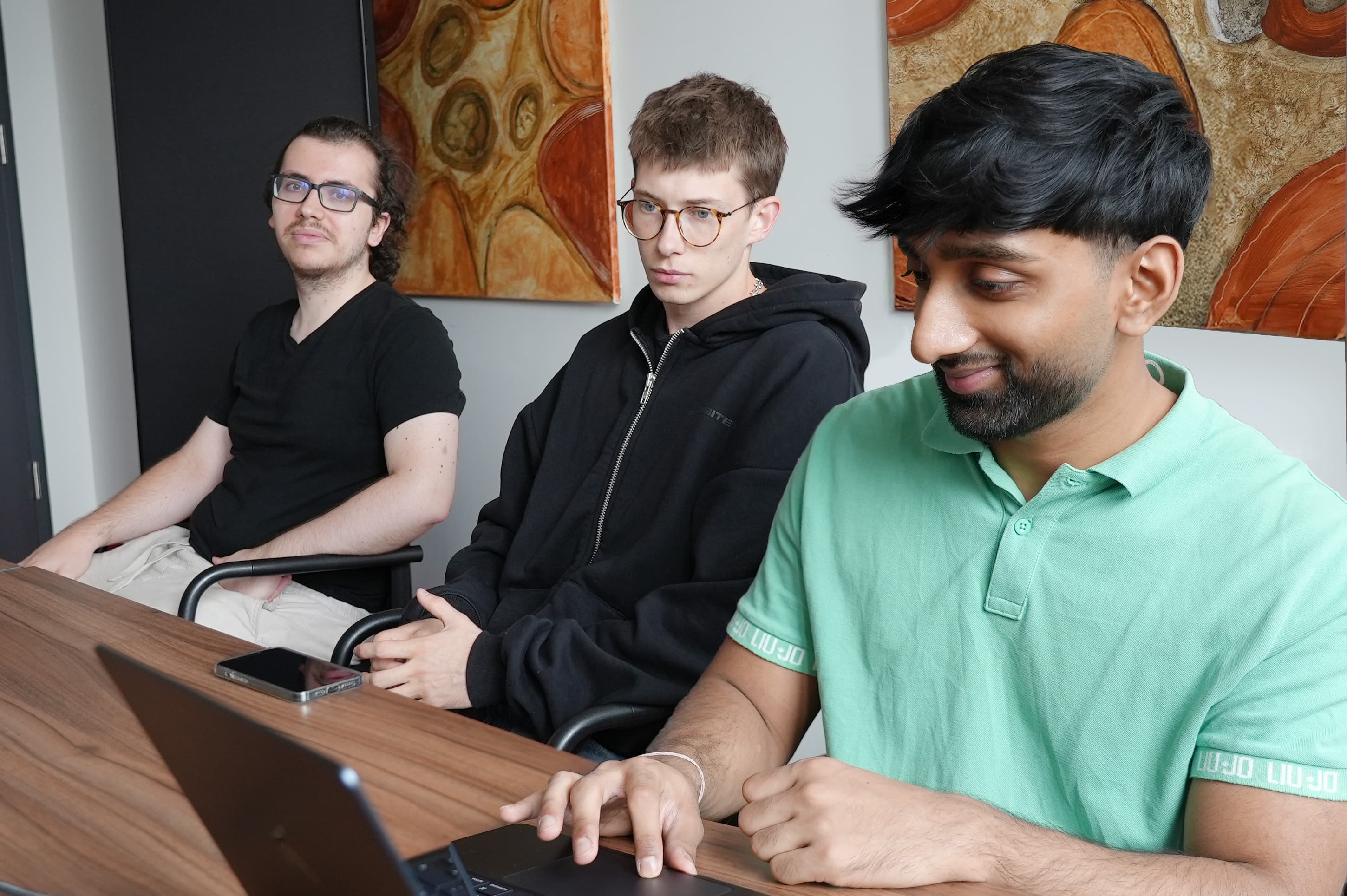 Drei Männer sitzen drinnen an einem Tisch. Zwei blicken in die Kamera, während der dritte, der ein grünes Hemd trägt, auf einen Laptop schaut. An der Wand hinter ihnen hängen abstrakte Gemälde.