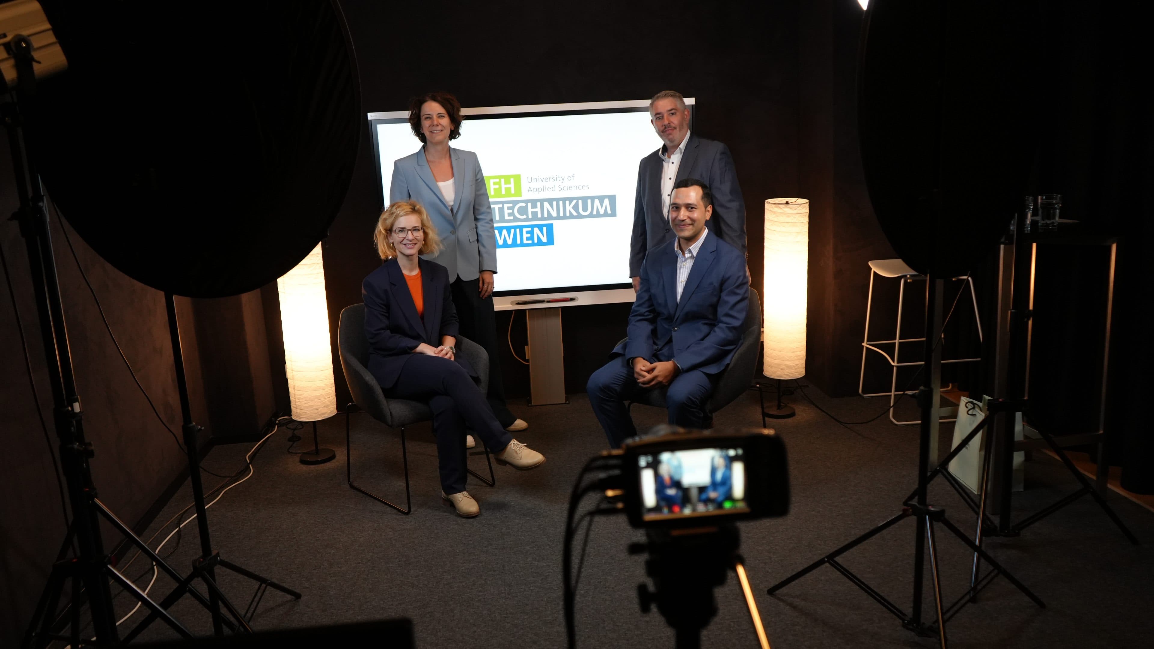 Vier Personen in Geschäftskleidung posieren in einem Studio mit Beleuchtung, einer Kamera und einer Leinwand, auf der „FH Technikum Wien“ zu sehen ist. Zwei sitzen, zwei stehen dahinter, alle lächeln. Die Kulisse wirkt professionell und modern.