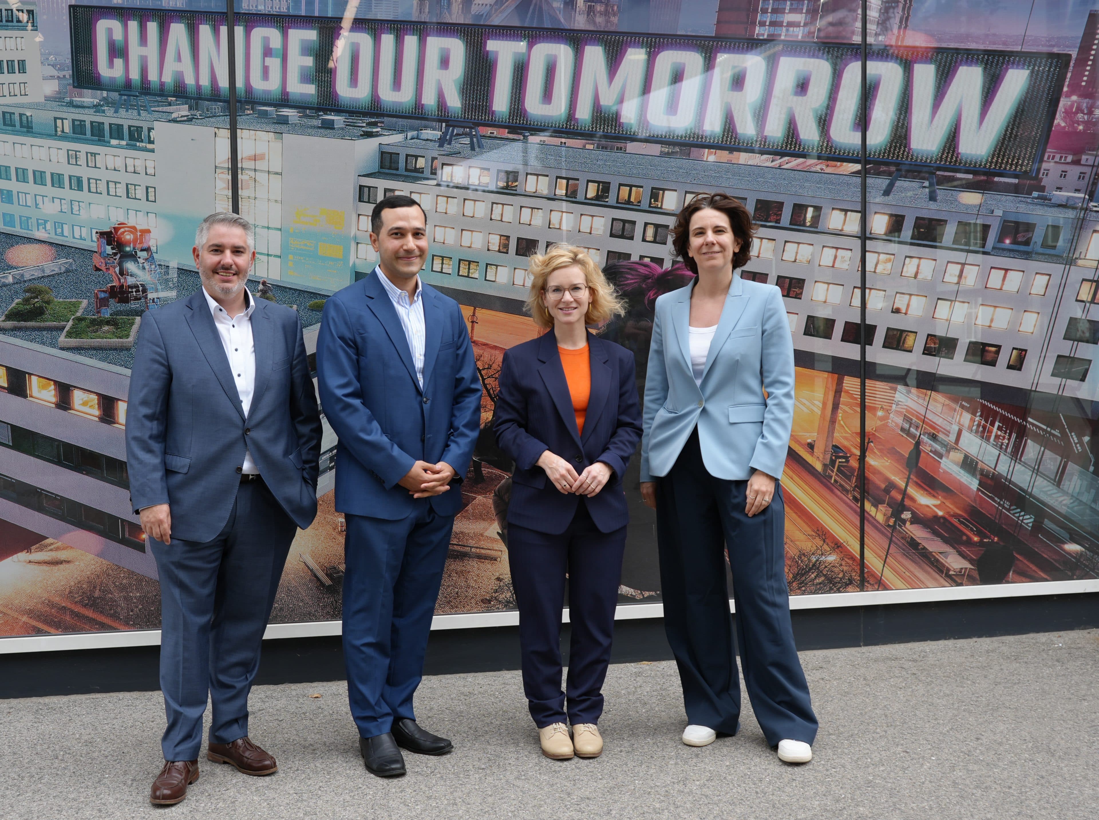 Vier Personen in Geschäftskleidung stehen draußen vor einem Wandbild mit der Aufschrift „CHANGE OUR TOMORROW“, lächeln und posieren für ein Gruppenfoto auf einer Straße in der Stadt.