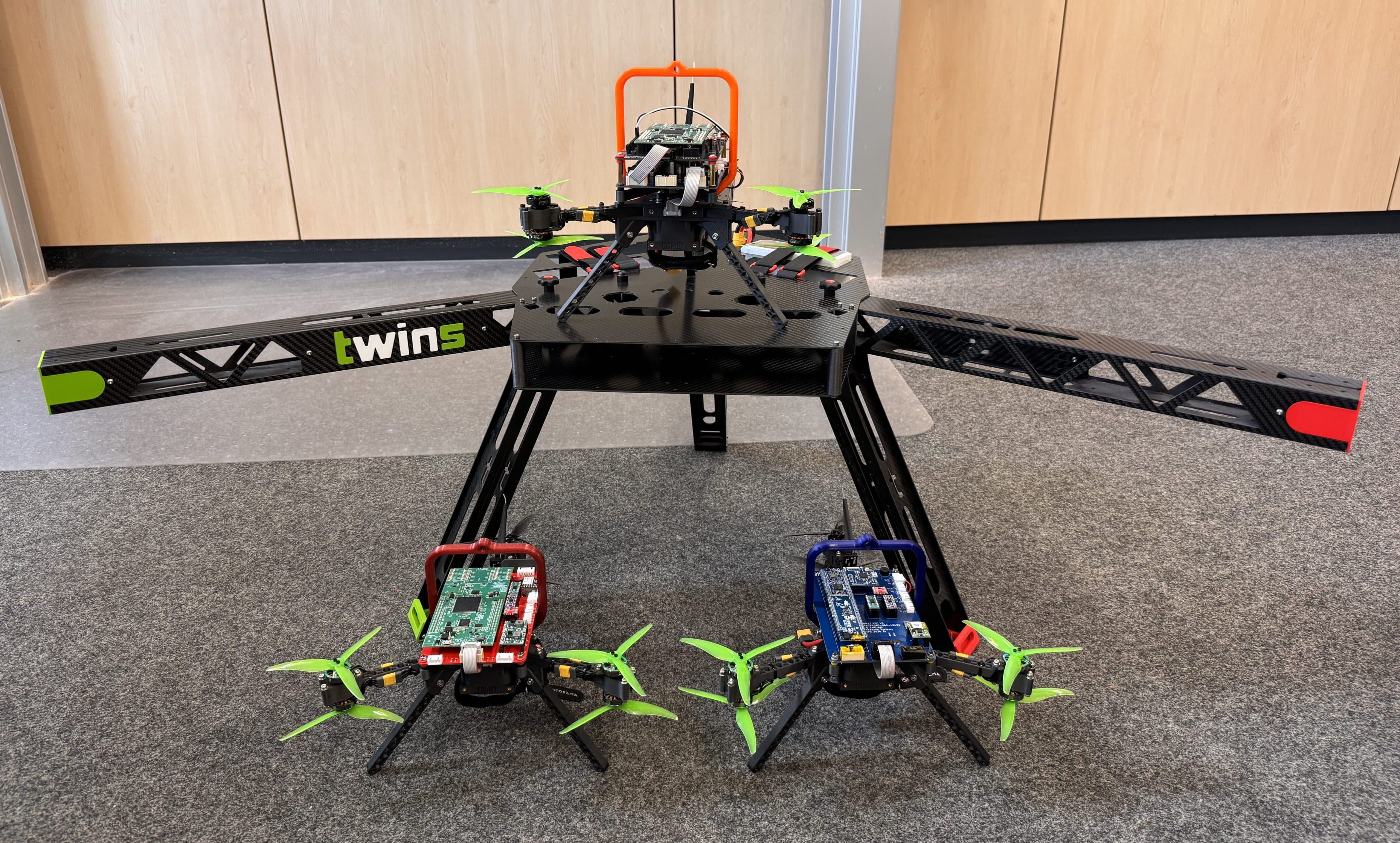 Auf dem Boden steht eine große schwarze Quadcopter-Drohne mit zwei Propellern an jedem Arm. Vor ihr stehen zwei kleinere Drohnen mit grünen Propellern und Leiterplatten – alles in einem Innenraum auf einem grauen Teppich.